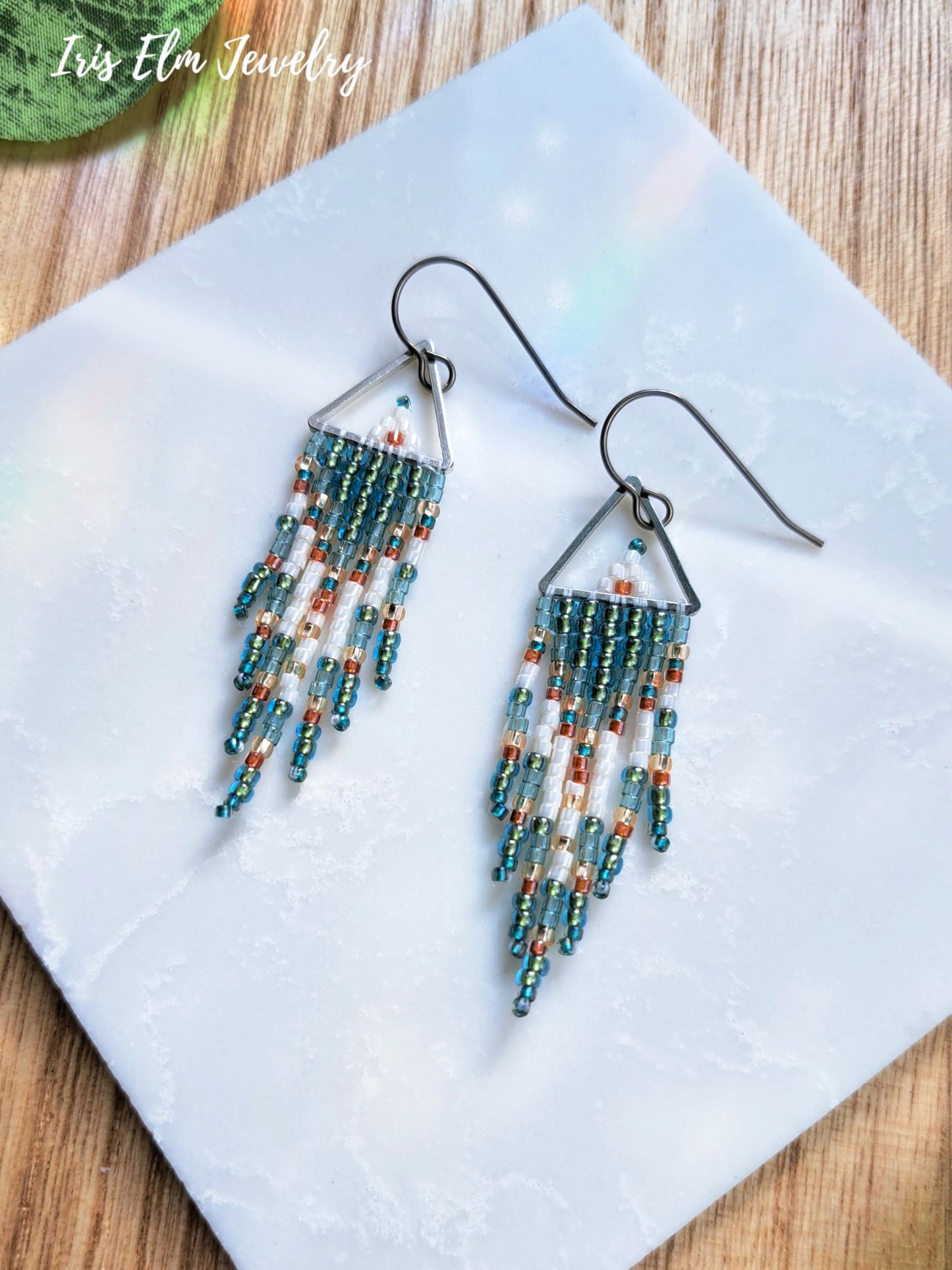 A pair of dangling beaded earrings with triangular silver-toned tops and multiple fringe strands in teal, white, and orange, displayed on a white square tile over a wood surface. The text 'Iris Elm Jewelry' appears in the top left corner.