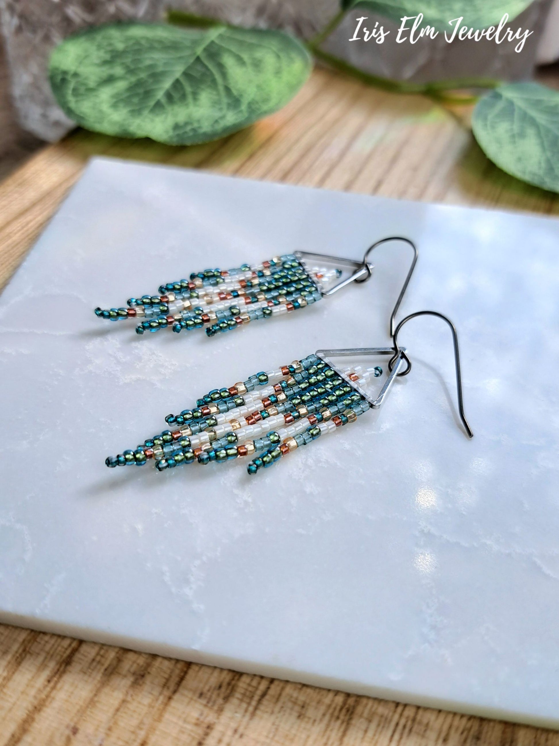 A pair of beaded dangle earrings with teal, white, and copper-colored beads on silver-toned fishhook wires, displayed on a white marble tray with green leaves in the background. The text 'Iris Elm Jewelry' is visible in the top right corner.