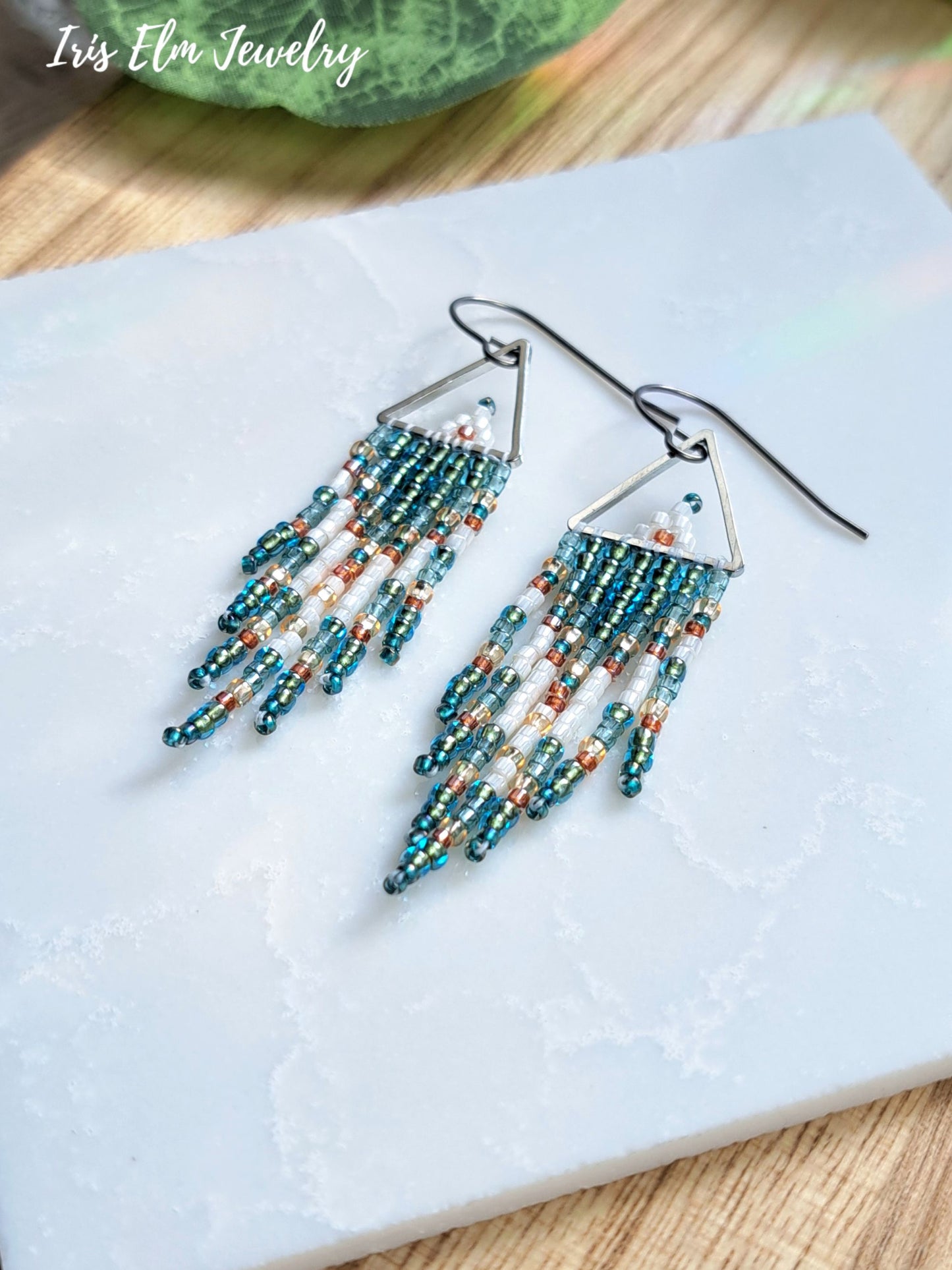 A pair of dangling beaded earrings with silver-toned triangular tops and long fringe made of teal, blue, white, and peach beads, displayed on a white stone slab. The text 'Iris Elm Jewelry' appears in the top left corner.
