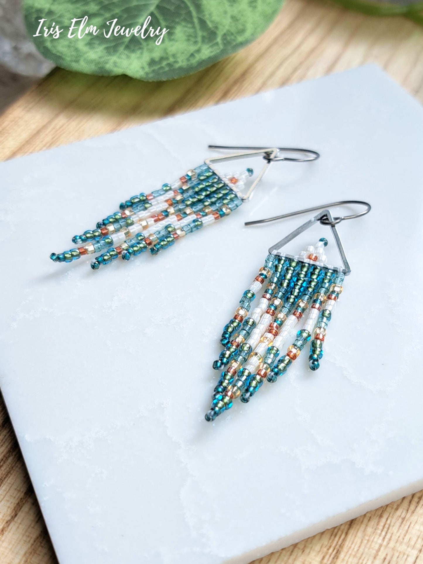 A pair of beaded dangle earrings with silver fishhook backs, featuring teal, white, and orange seed beads arranged in layered fringe. The earrings are displayed on a white textured square tile, with a wooden surface and a green leaf in the background.