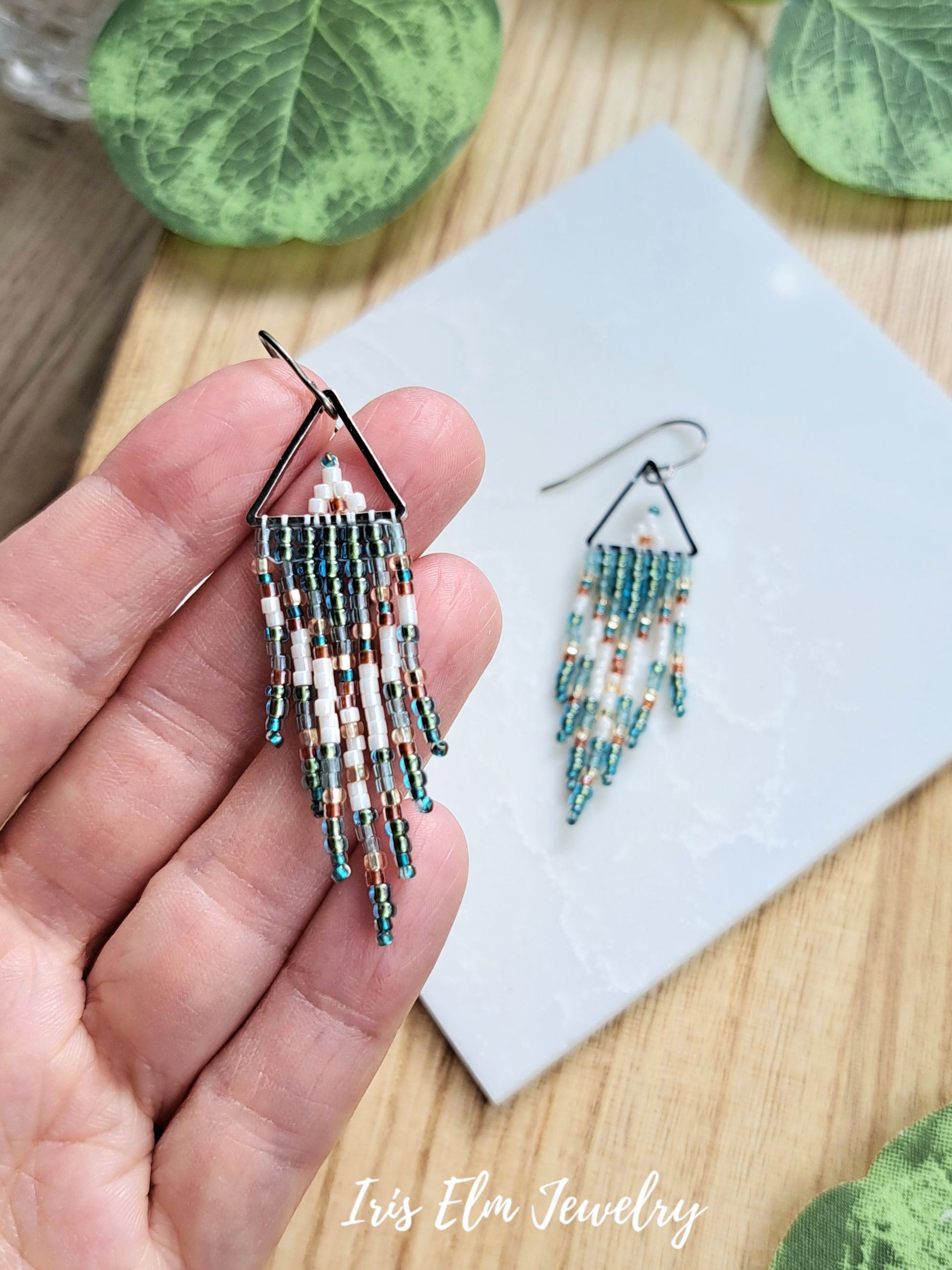 A hand holds a pair of dangling beaded earrings with a geometric triangular top and long fringe, next to a white display card on a wooden surface with green leaves.