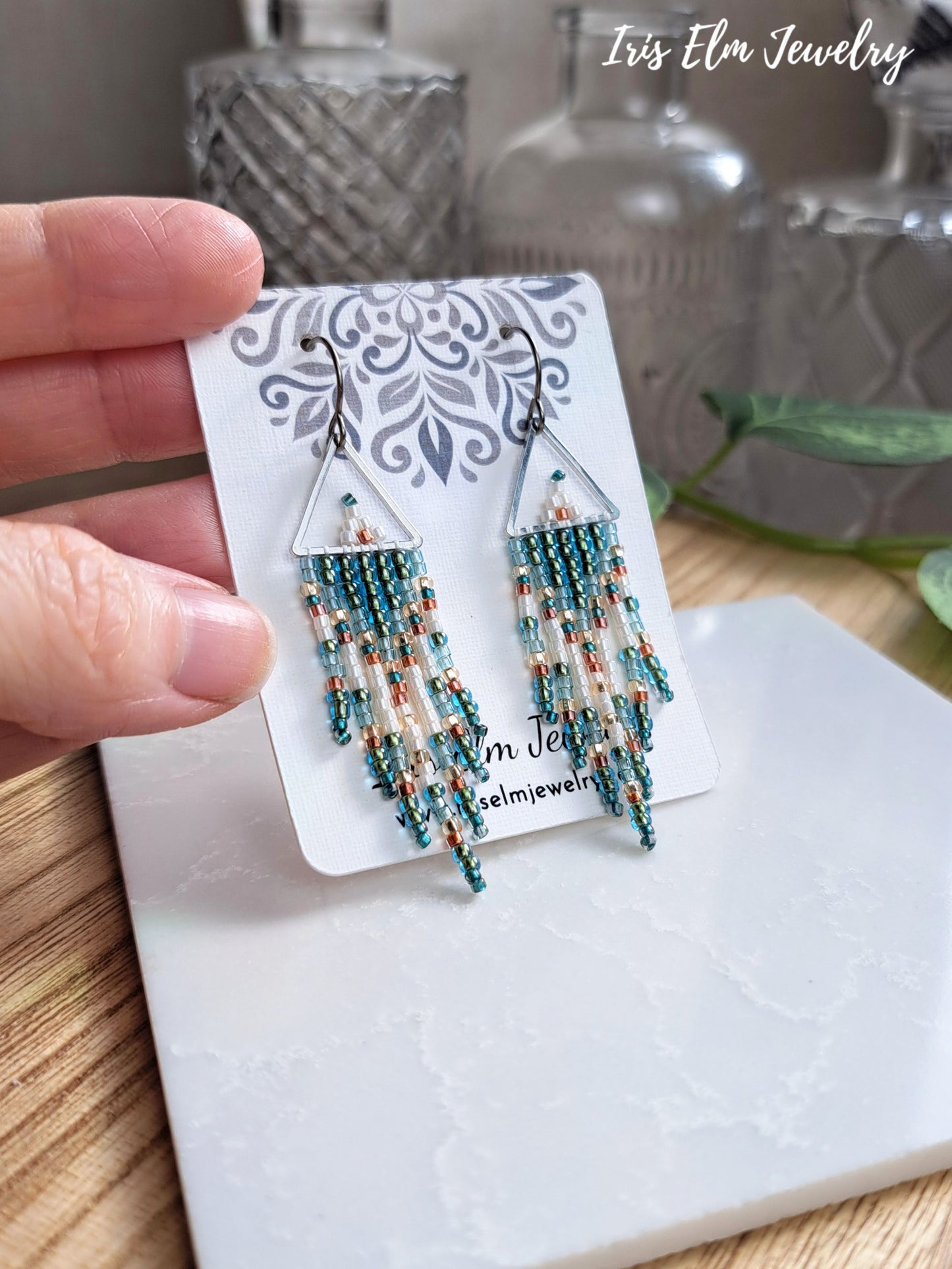 A hand holds a pair of dangling beaded earrings on a branded display card. The earrings have triangular clear frame tops with multiple strands of turquoise, teal, and peach beads hanging below.