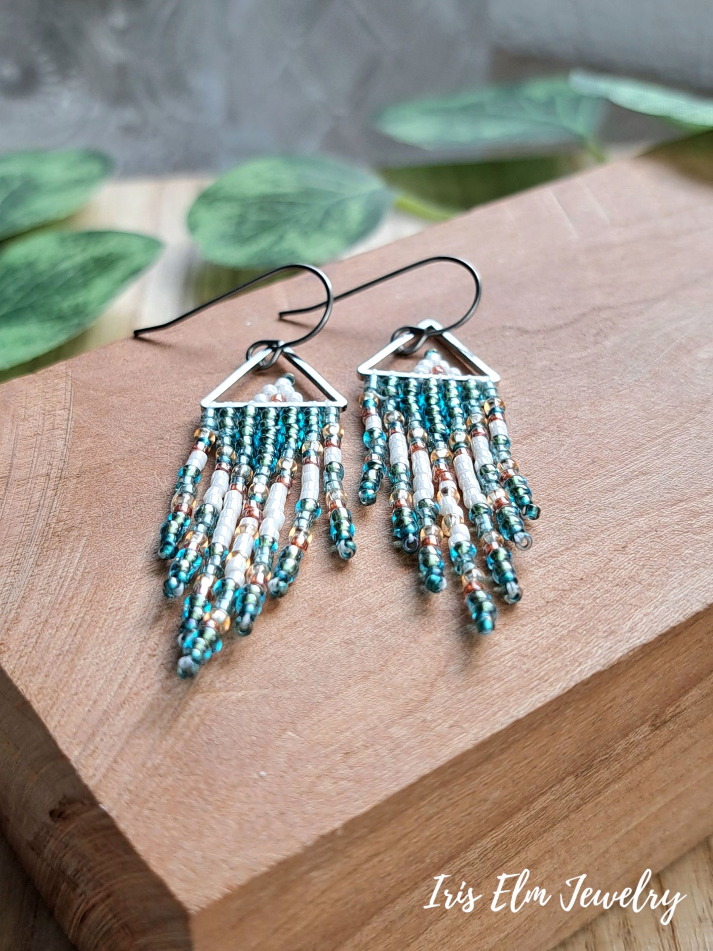 A pair of dangling beaded earrings with triangular silver-toned tops and multiple fringe strands, displayed on a wooden block with green leaves in the background. The brand name 'Iris Elm Jewelry' is visible in the bottom right corner.