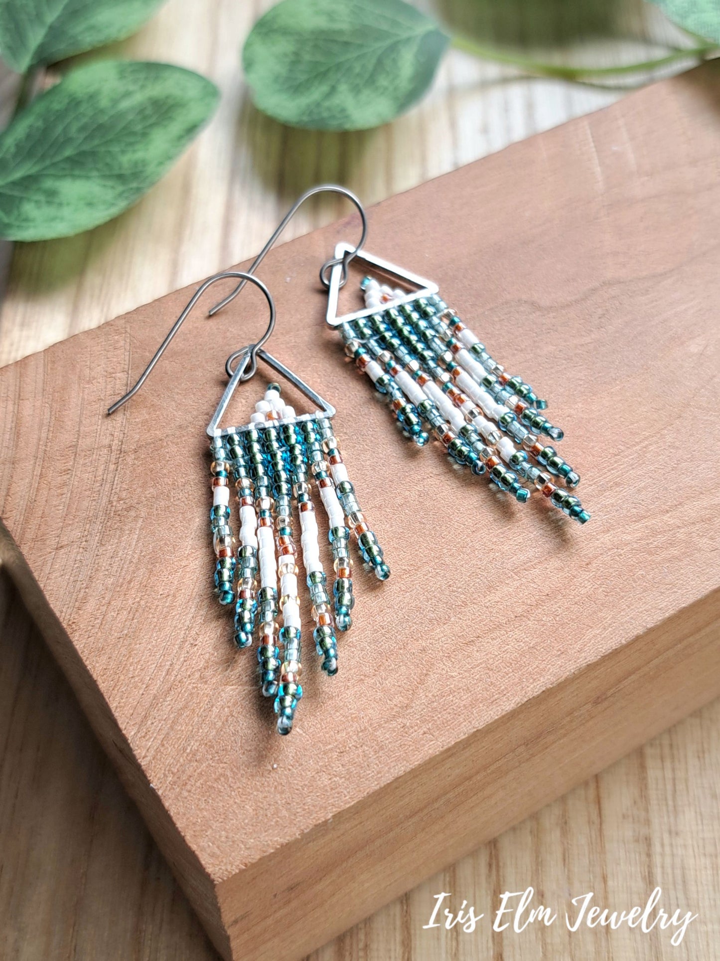 A pair of bohemian-style beaded fringe dangle earrings with silver hooks, displayed on a wooden block with green leaves in the background. The brand name 'Iris Elm Jewelry' is visible in the bottom right corner.
