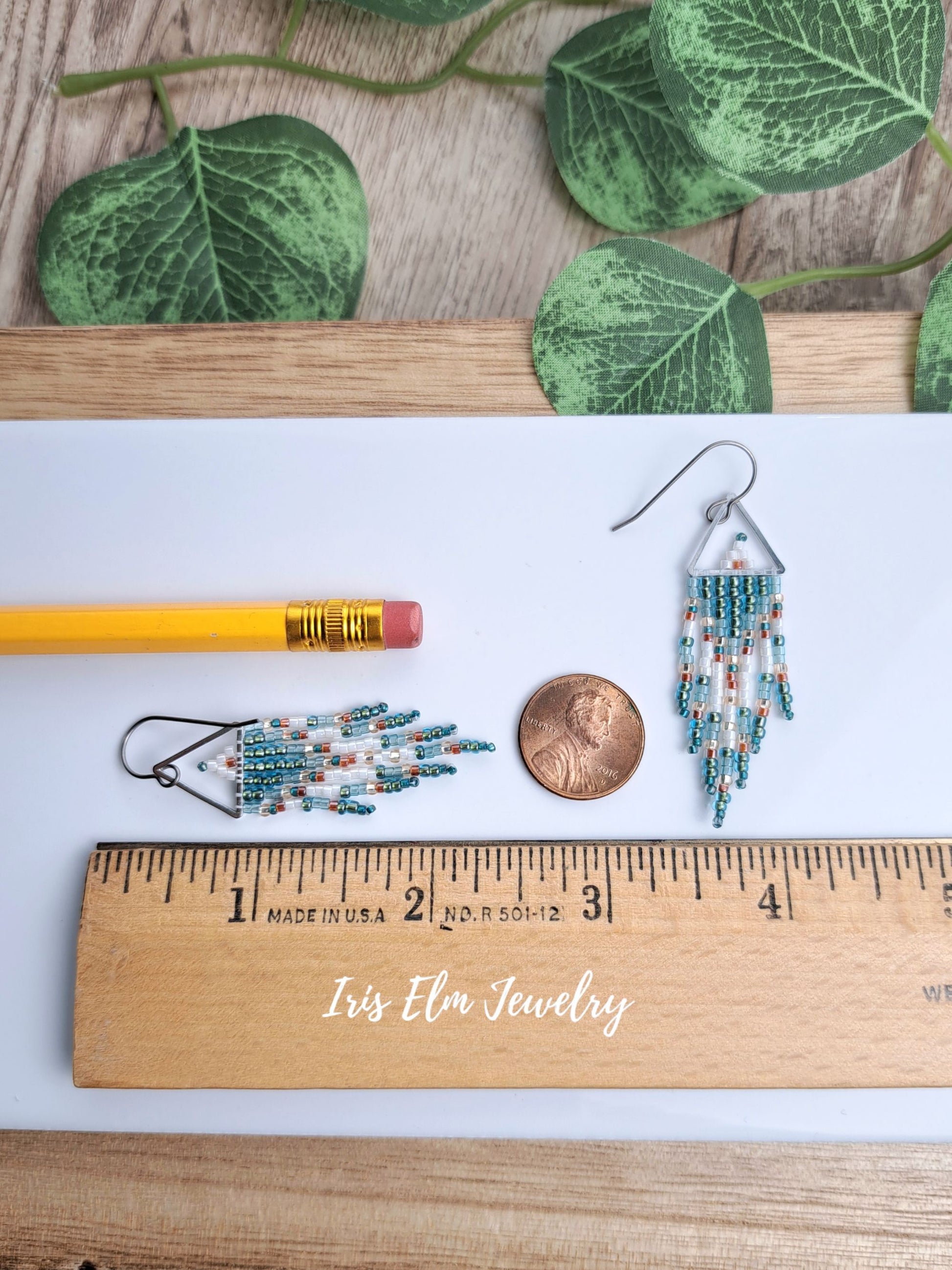 Two handmade 2-3/8 inch long beaded dangle earrings displayed on a white surface with a yellow pencil, a penny, a wooden ruler, and green leaves in the background. The ruler is branded 'Iris Elm Jewelry'.