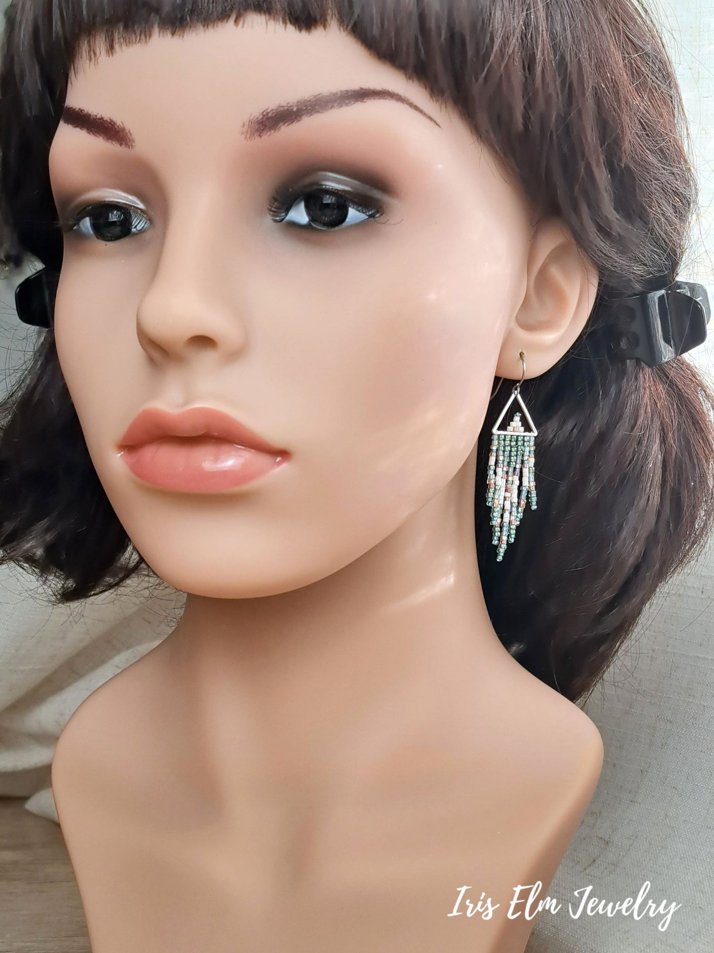 A mannequin bust with short dark brown hair and a black hair clip, wearing a dangling silver earring with a triangular top and fringe drops. The mannequin is photographed against a light neutral background.
