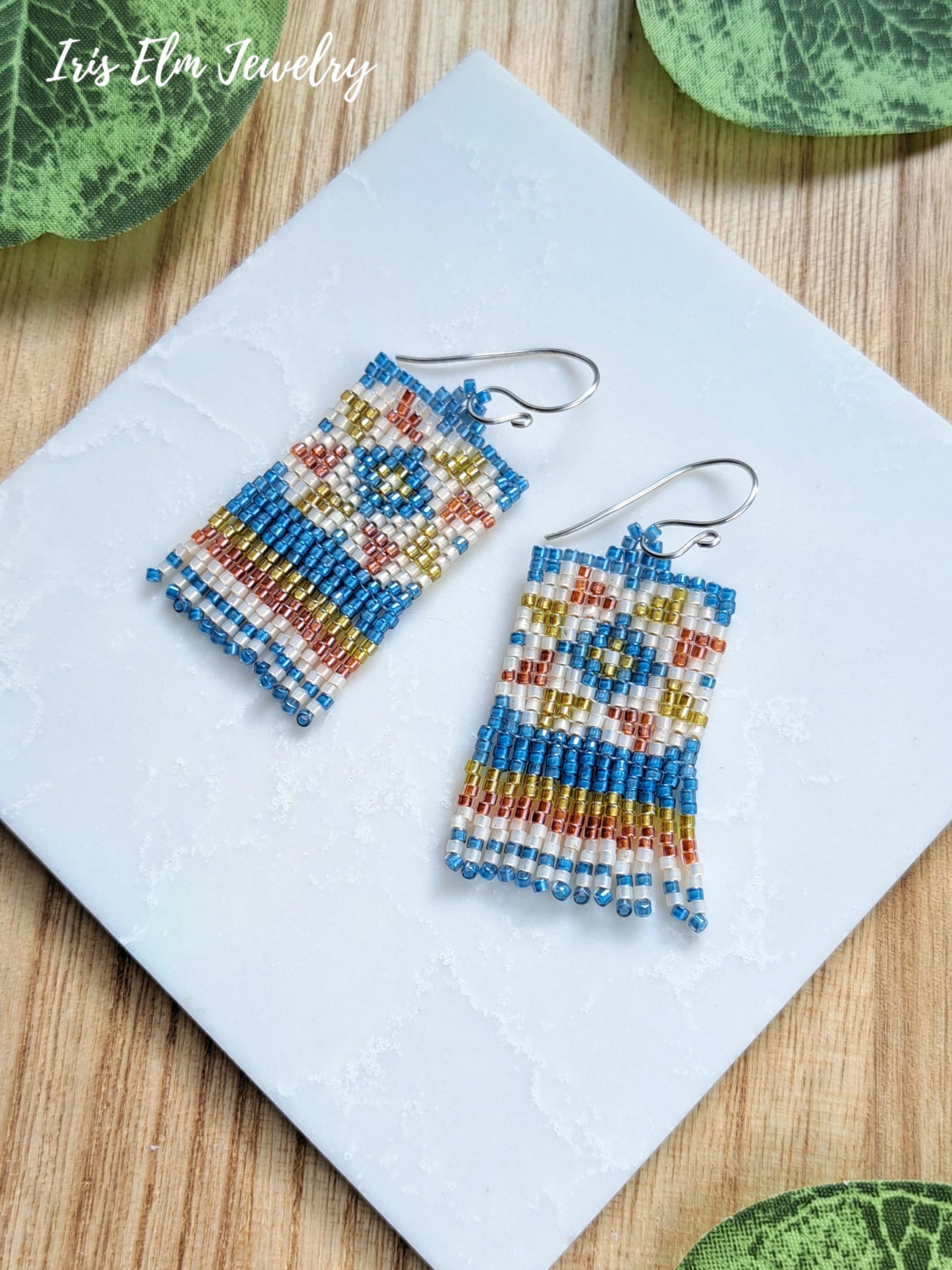 A pair of handmade beaded dangle earrings with geometric quilt patterns in blue, cream, yellow, and orange, hanging from silver fishhook wires on a white textured square card over a wooden surface.