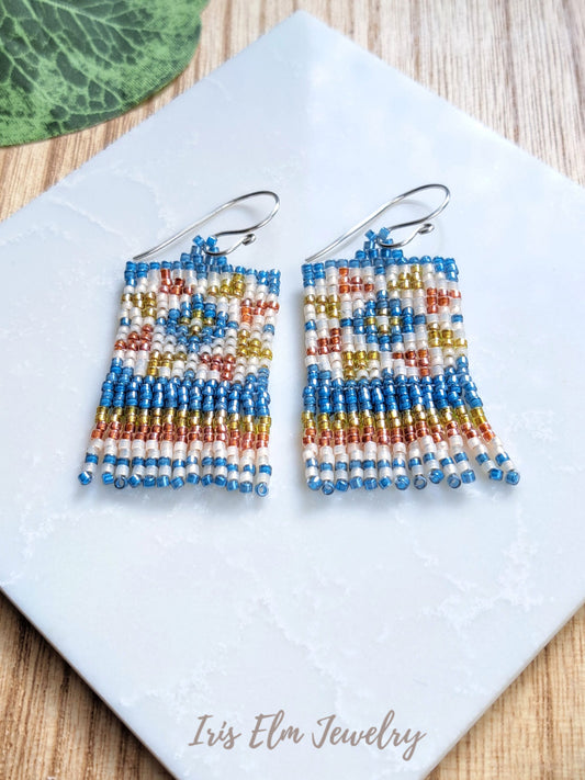 Handmade beaded dangle earrings with a geometric quilt block pattern in blue, white, orange, and gold, hanging from silver fishhook wires on a white ceramic tile.