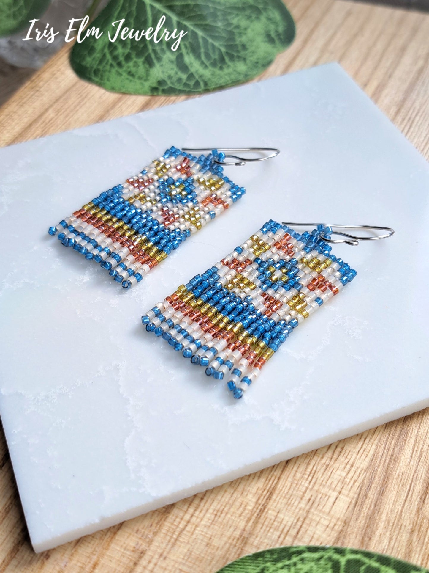 A pair of rectangular beaded drop earrings with silver fishhook wires, featuring intricate geometric quilt block patterns in blue, cream, yellow, and orange. The earrings are displayed on a white textured tile over a light wood surface, with the brand name 'Iris Elm Jewelry' visible in the top left corner.