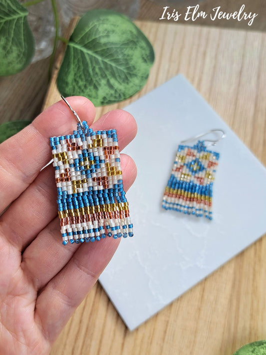 A hand holds a small rectangular beaded fringe seed beadearring with a sterling silver fishhook ear wire. Another matching beaded earring rests on a white card on a light wood surface, with green leaves in the background.