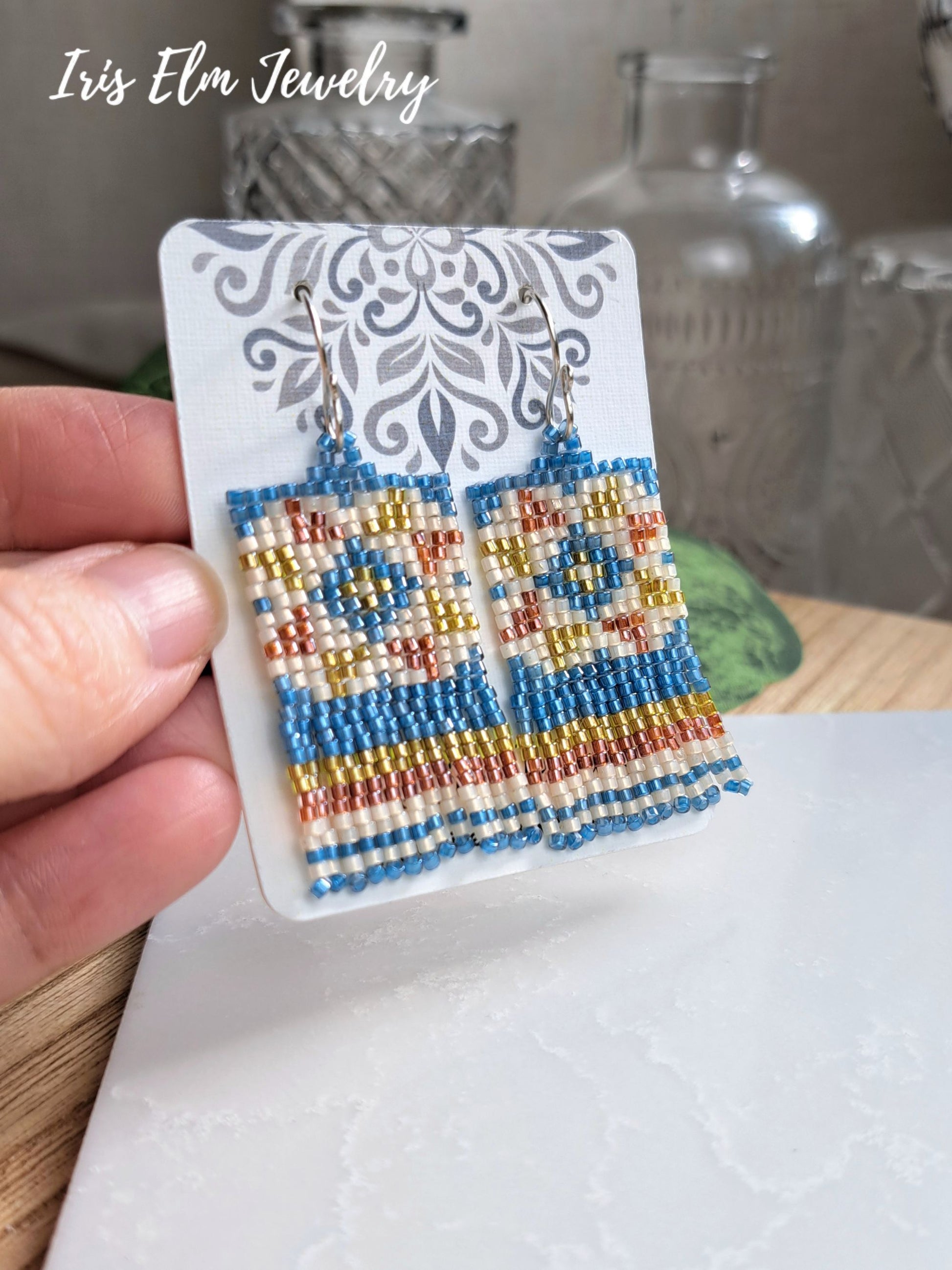 Hand holding a display card with a pair of rectangular beaded dangle earrings. The earrings feature a Southwestern-style geometric quilt pattern in blue, yellow, cream, and terracotta red, suspended from silver fishhook wires.