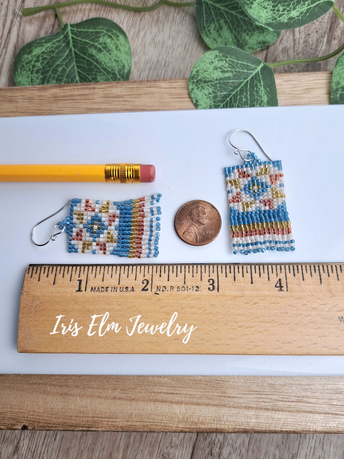 Two small 2 inch long beaded drop earrings with a geometric pattern in blue, white, orange, and yellow, displayed on a white sheet of paper next to a pencil, a penny, and a wooden ruler. The text 'Iris Elm Jewelry' is written on the ruler.