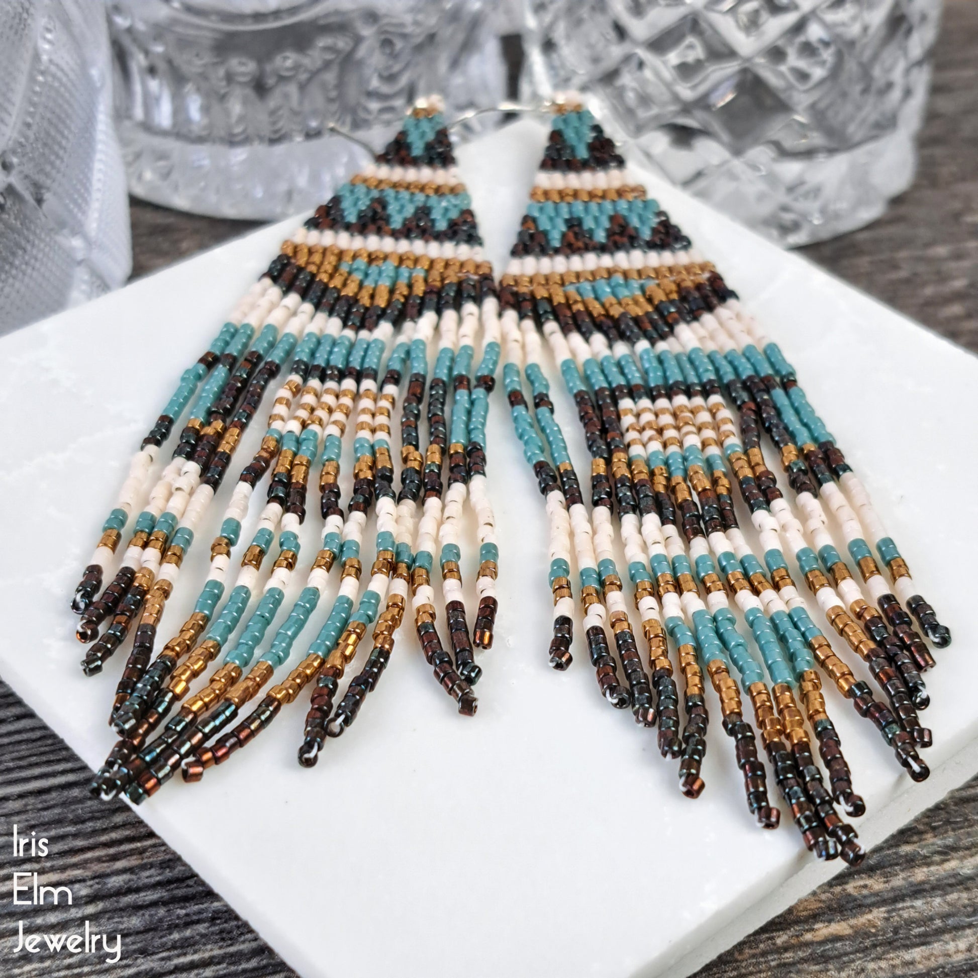 Beaded long fringe earrings made with blue, brown, bronze and cream glass seed beads on a white plate with a blurred background