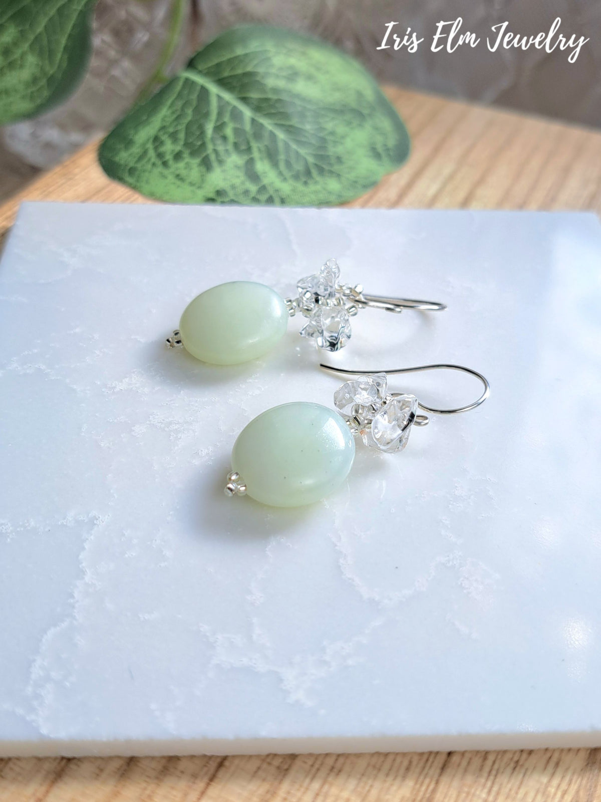 A pair of dangle earrings by Iris Elm Jewelry, featuring pale green oval beads and clear crystal flower clusters on silver hooks, displayed on a white marble slab.