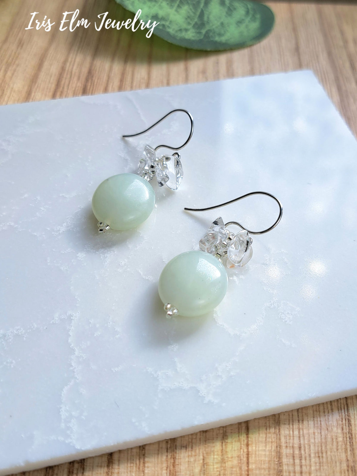 A pair of dangle earrings with pale green oval serpentine jade beads, clear faceted crystal accents, and silver-tone fishhook wires, displayed on a white marble slab over a wood surface. The text 'Iris Elm Jewelry' appears in the upper left.