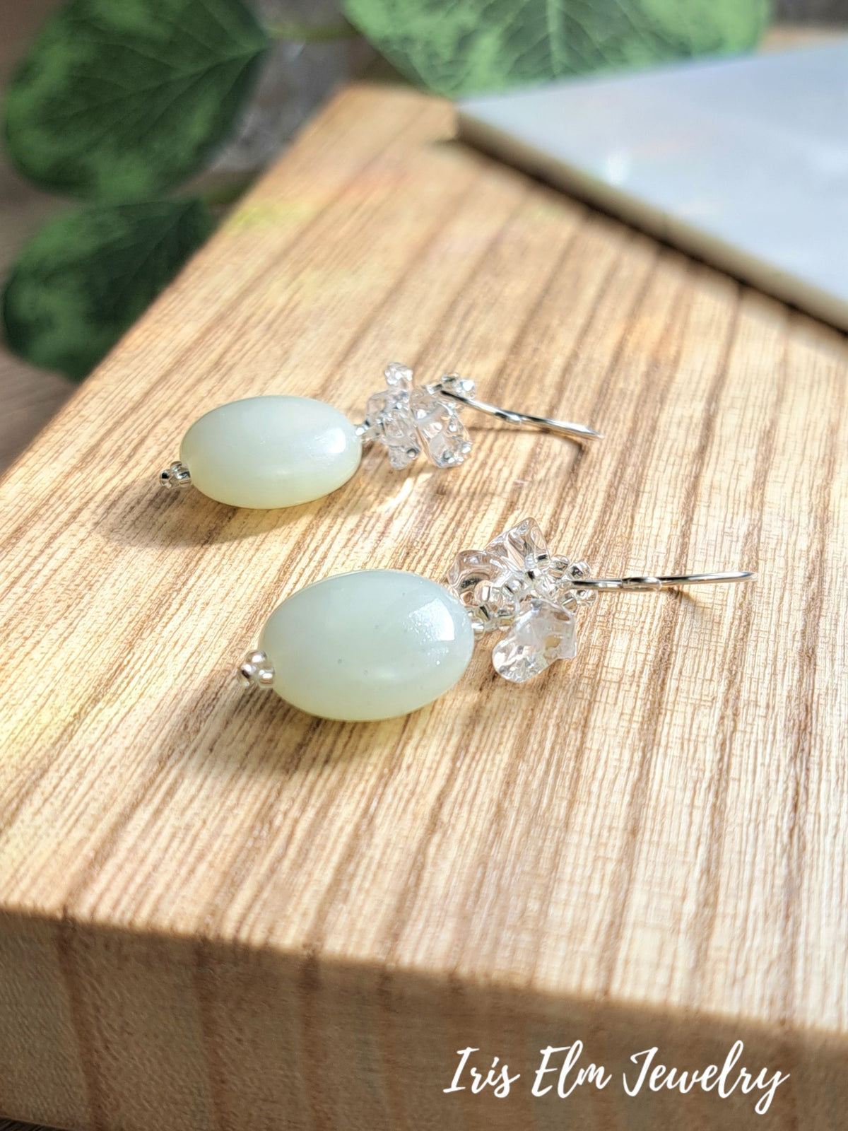 A pair of dangle earrings with oval pale green serpentine jade beads and clear crystal floral clusters on silver hooks, displayed on a wooden block with a blurred green leaf and white object in the background. The text 'Iris Elm Jewelry' is visible in the bottom right corner.
