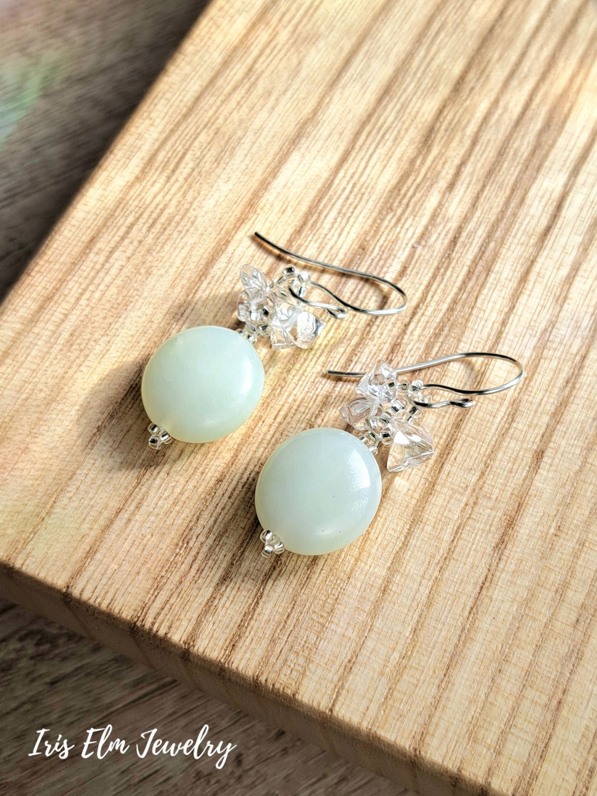 A pair of dangle earrings with round pale green serpentine jade beads, clear crystal floral clusters, and silver fishhook wires, displayed on a light wood surface. The brand name 'Iris Elm Jewelry' is visible in the bottom-left corner.