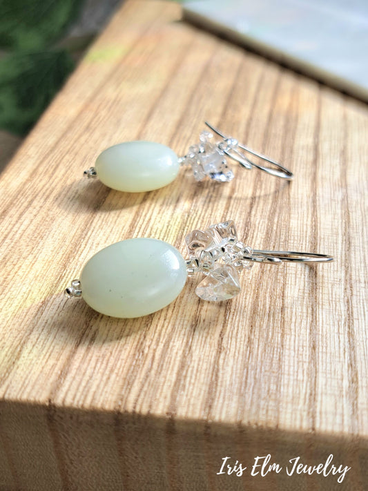 A pair of dangle earrings with oval milky green serpentine jade beads and clear faceted crystal accents on silver hooks, displayed on a wooden block with the brand name 'Iris Elm Jewelry' visible in the bottom right corner.