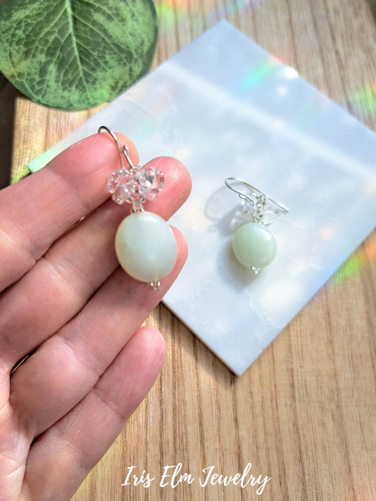 A hand holds a pair of dangling earrings with pale green round beads, clear crystal-like floral tops, and silver hooks, with a second earring resting on a white card on a wooden surface.