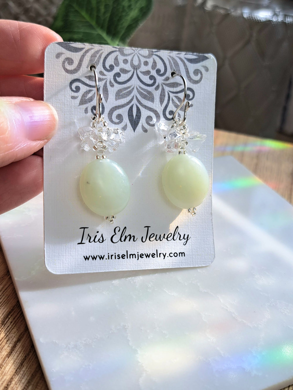A pair of dangle earrings with pale yellow-green oval serpentine jade beads, clear crystal quartz stone accents, and silver-toned hooks, displayed on a white card labeled Iris Elm Jewelry, held by a hand.