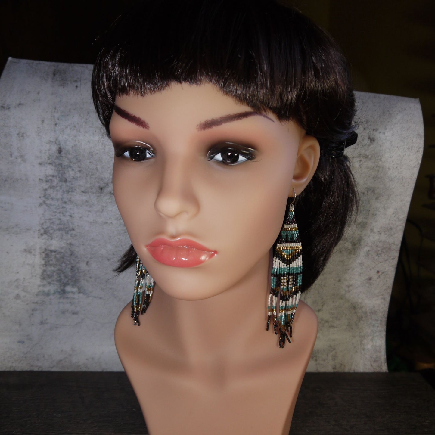 Mannequin head with dark hair and beaded long fringe earrings on a textured background