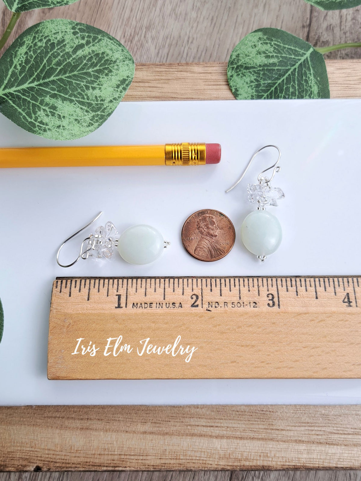 A pair of 1-5/8 inch long dangle earrings with pale green oval beads and silver hooks are displayed on a white sheet with a pencil, penny, wooden ruler, and green leaves. The brand name 'Iris Elm Jewelry' is printed on the ruler.