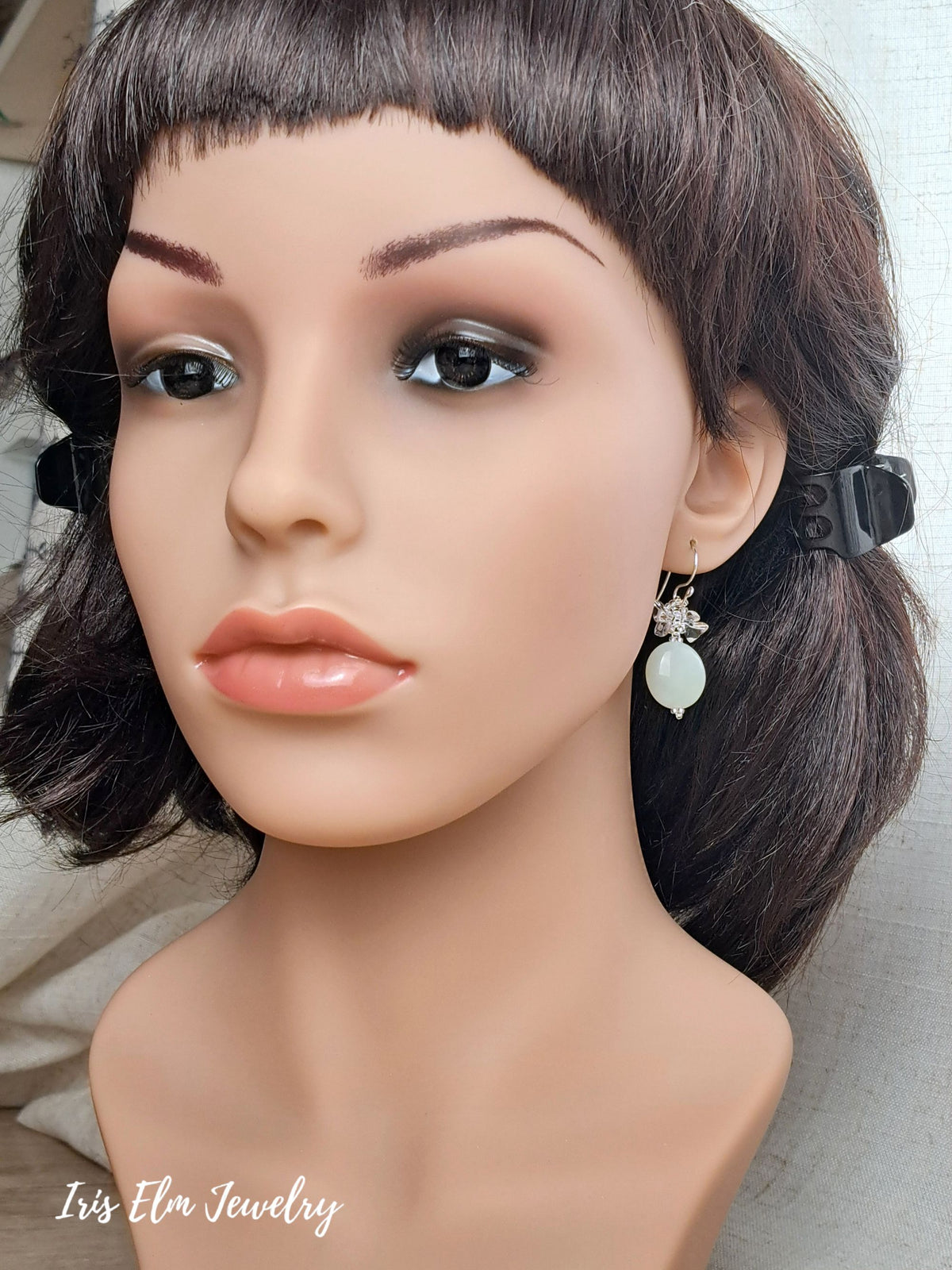 A close-up of a mannequin bust wearing a dangling serpentine jade earring. The mannequin has dark brown hair and a neutral beige background.