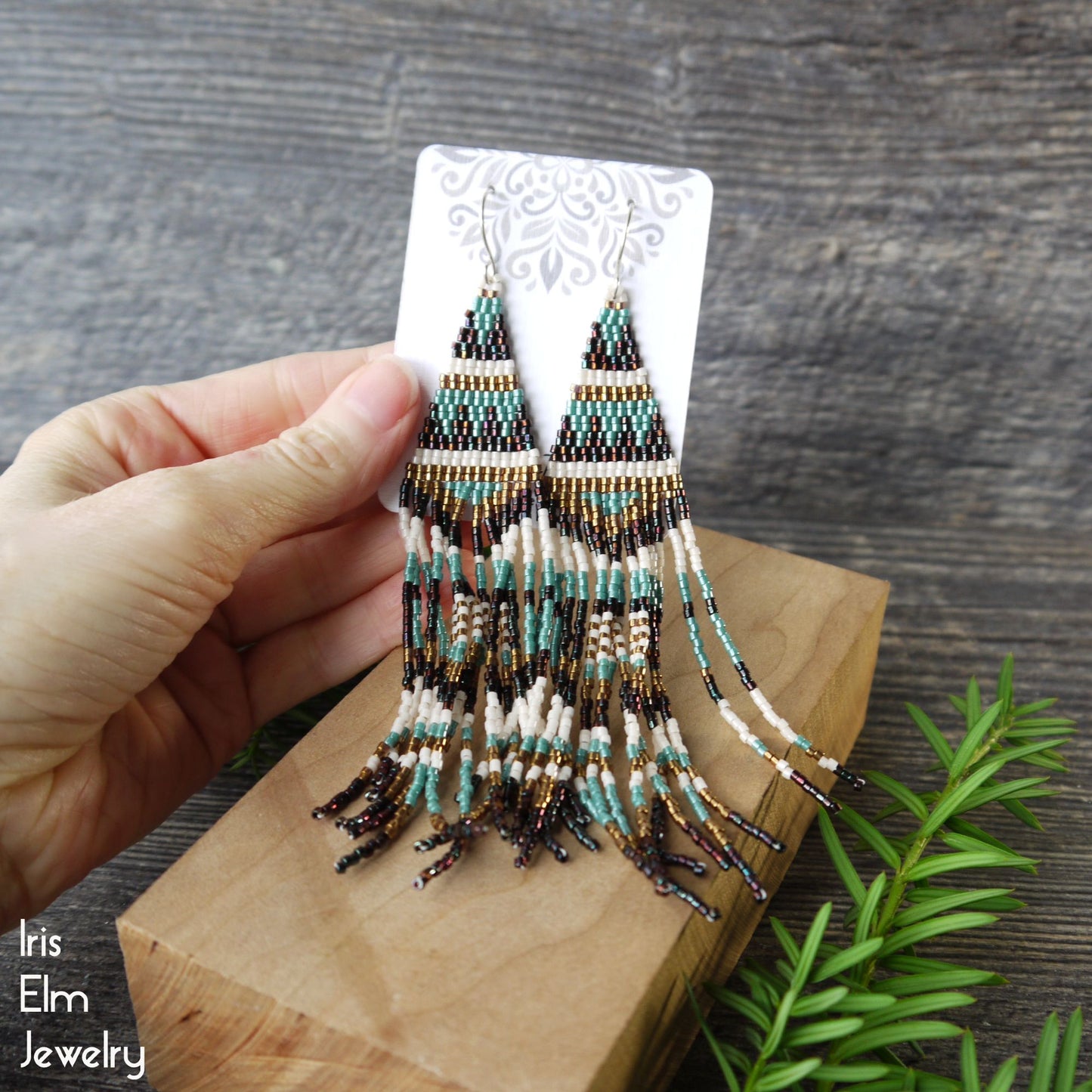 Hand holding beaded fringe bronze seed bead earrings on a wooden block with a natural background