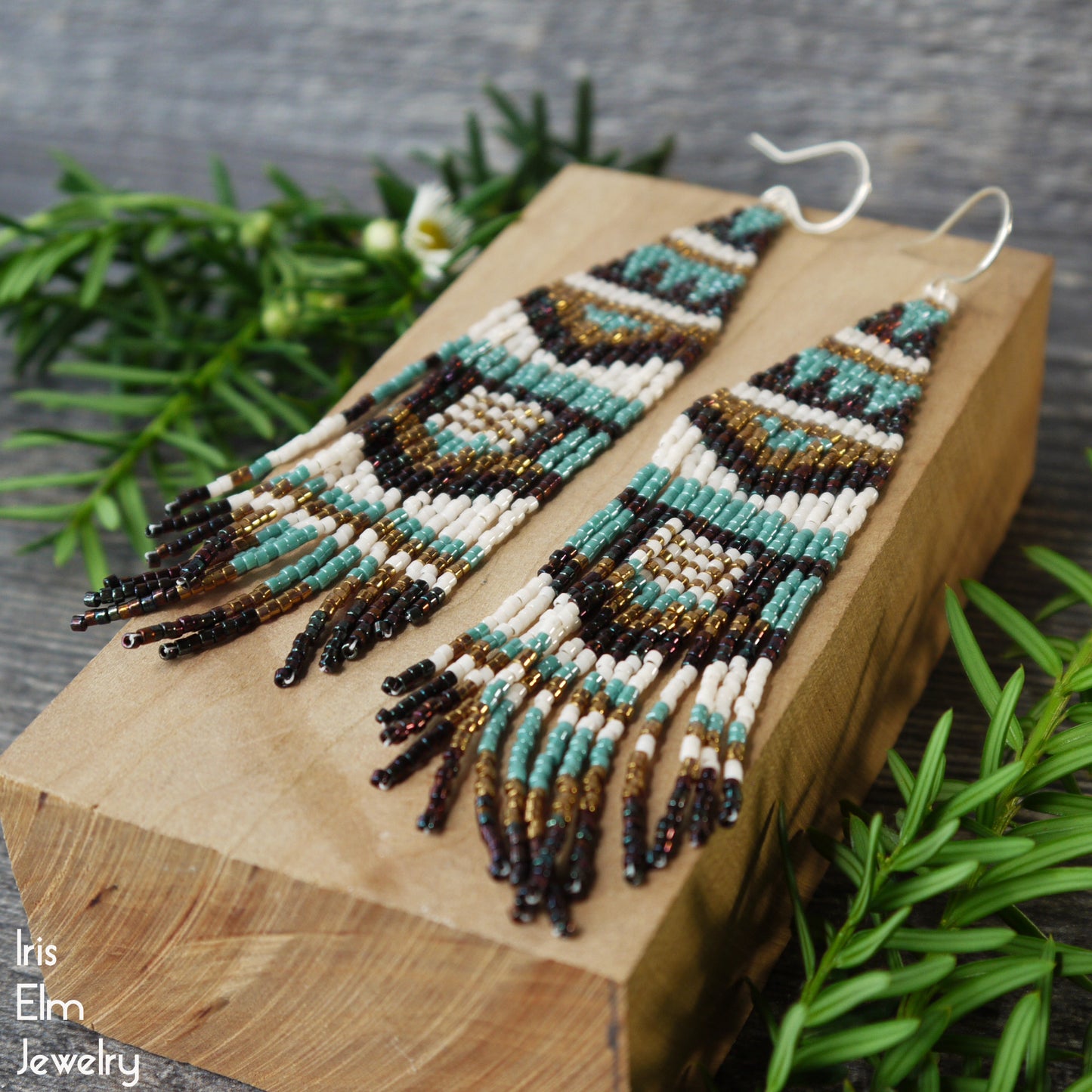 Long beaded seed bead brown and blue fringe earrings on a wooden block with greenery in the background, featuring the brand 'Iris Elm Jewelry'.