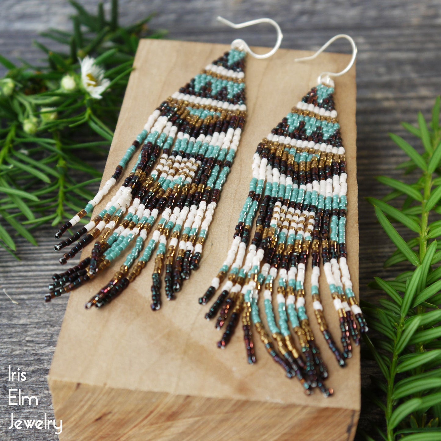 Beaded blue and bronze seed bead fringe earrings on a wooden block with greenery in the background, featuring the brand 'Iris Elm Jewelry'.