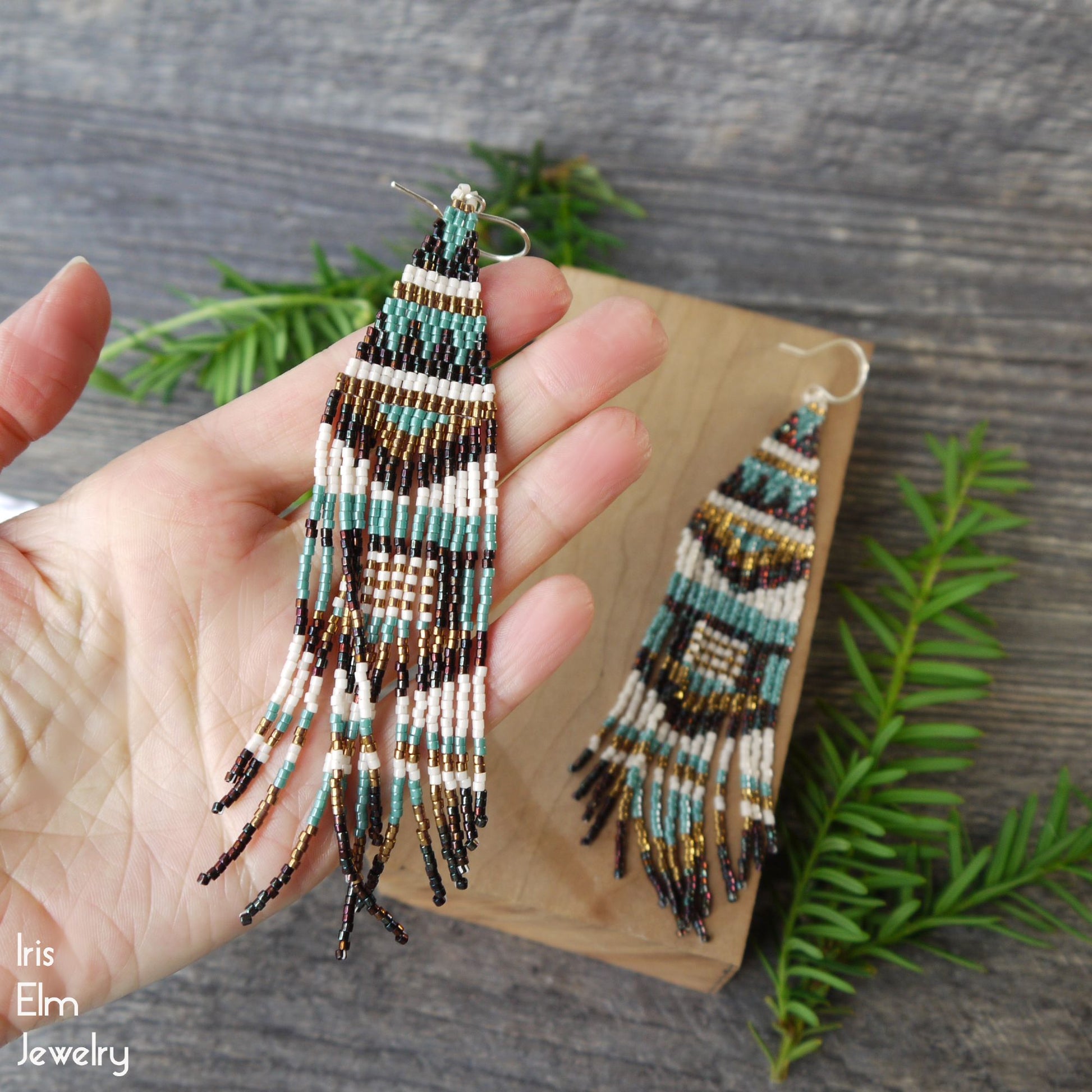 Beadwork seed bead fringe earrings held by a hand with a wooden display card and greenery in the background, featuring 'Iris Elm Jewelry' branding.