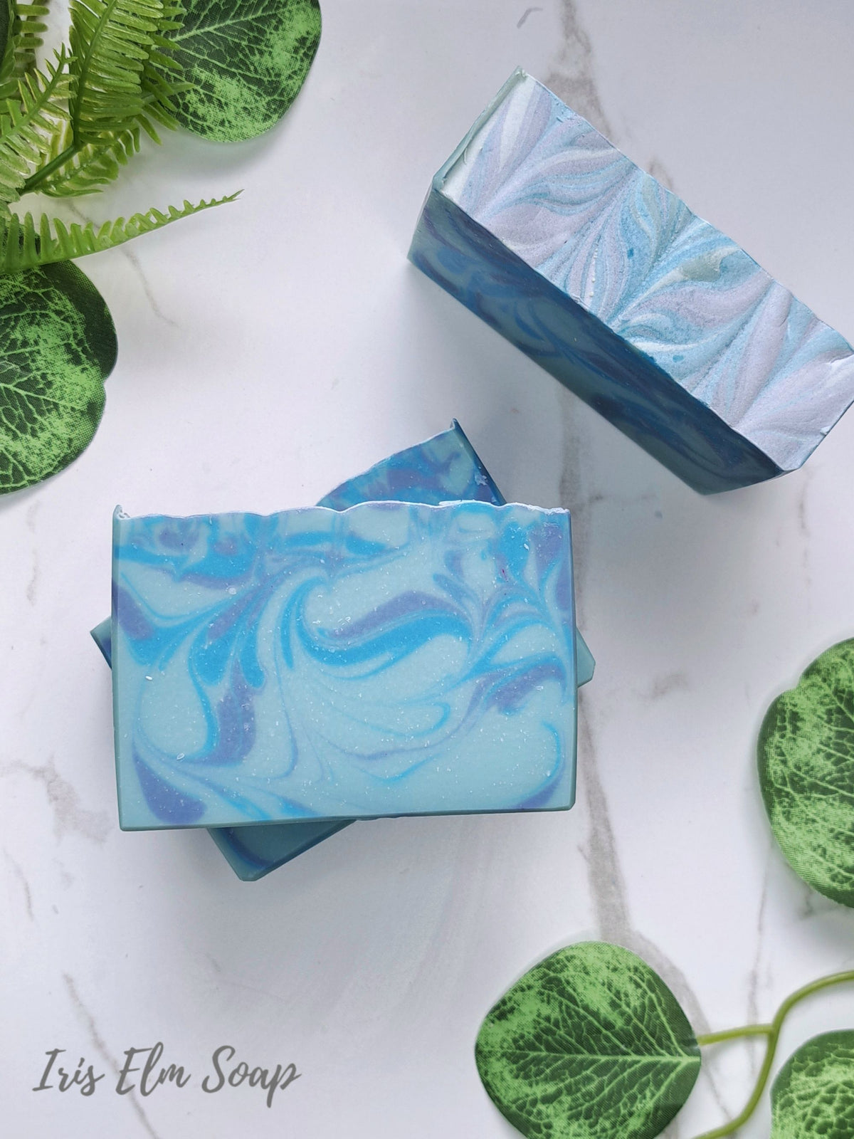 Two rectangular bars of blueberry Iris Elm Soap with a blue and white marbled swirl pattern, displayed on a white marble surface with green decorative leaves.