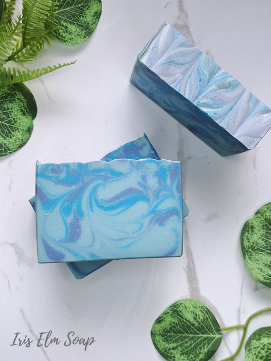 Two rectangular bars of blueberry Iris Elm Soap with a blue and white marbled swirl pattern, displayed on a white marble surface with green decorative leaves.