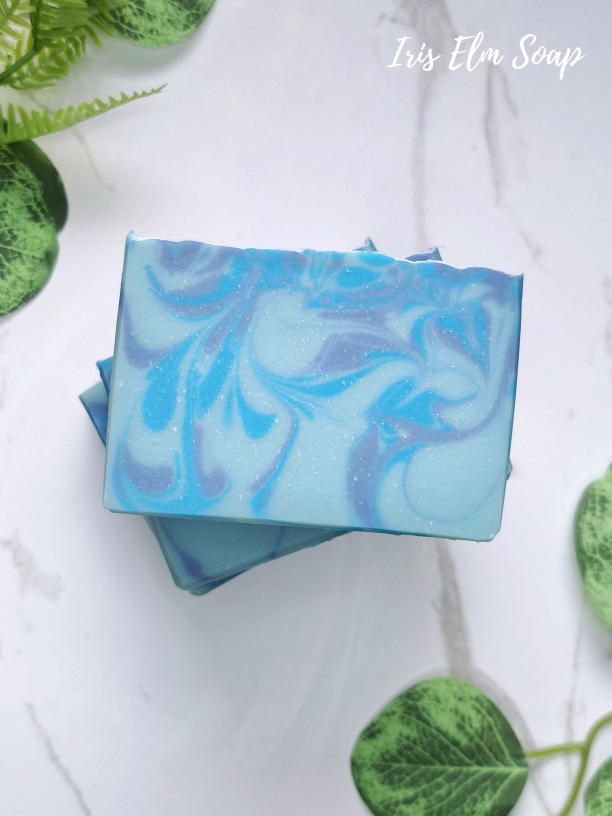 A rectangular bar of blueberry handmade soap with a light blue and blue marbled swirl pattern, stacked on another soap bar. The soaps are photographed on a white marble surface decorated with green leaves, with the text 'Iris Elm Soap' in the top right corner.