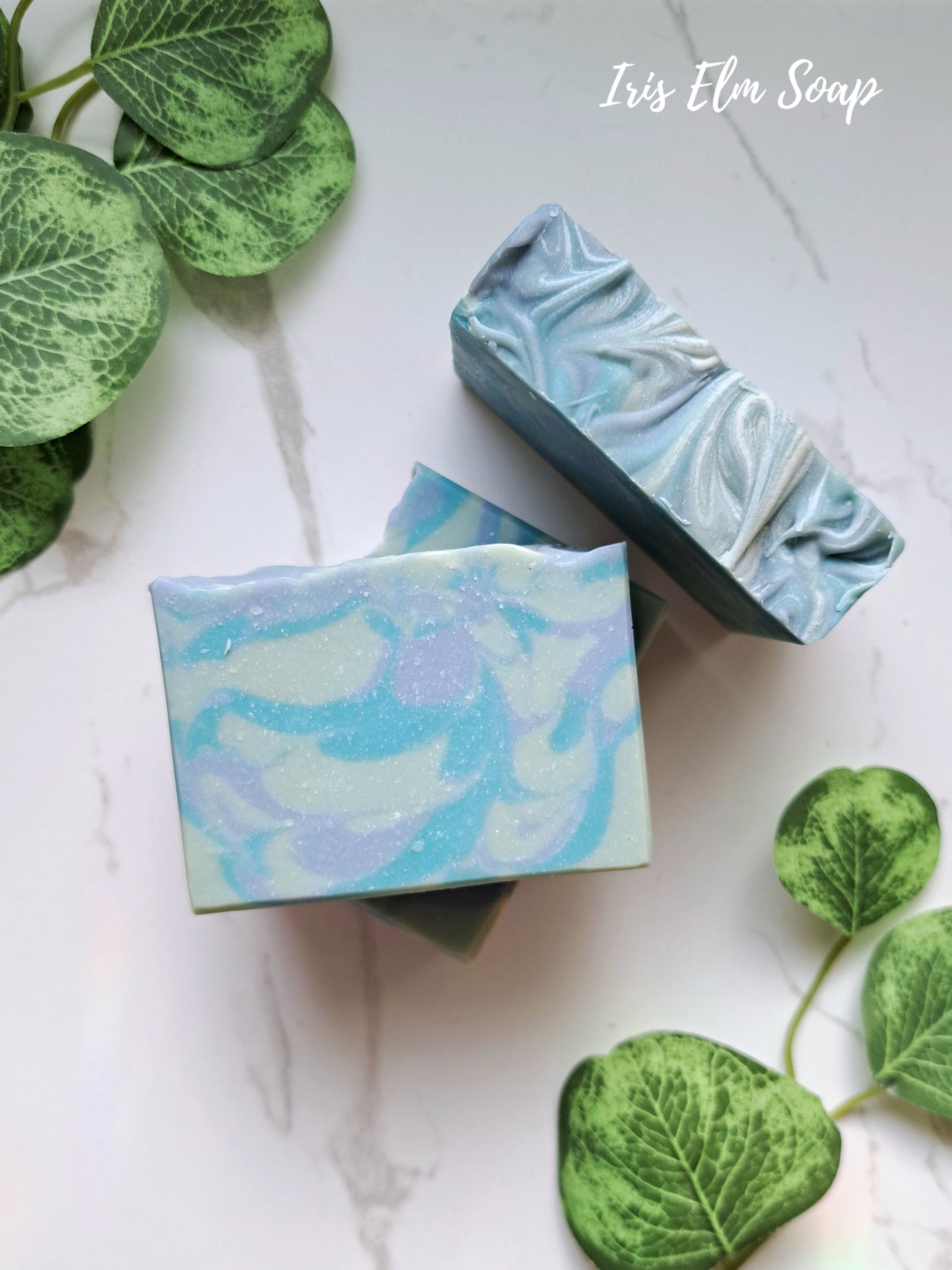Two bars of blueberry handmade soap with a marbled design on a white surface with green leaves.