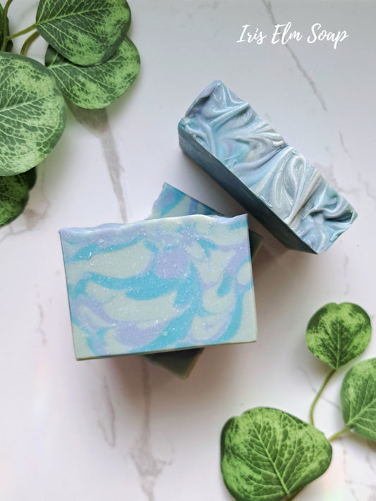 Two bars of blueberry handmade soap with a marbled design on a white surface with green leaves.