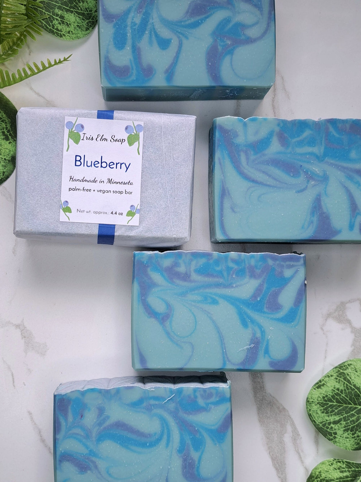 Five bars of artisan soap with blue and teal marbled swirls are arranged on a white marble surface alongside a wrapped package. The package features a white label with blue and green illustrations that read 'Iris Elm Soap' and 'Blueberry Handmade in Minnesota'.