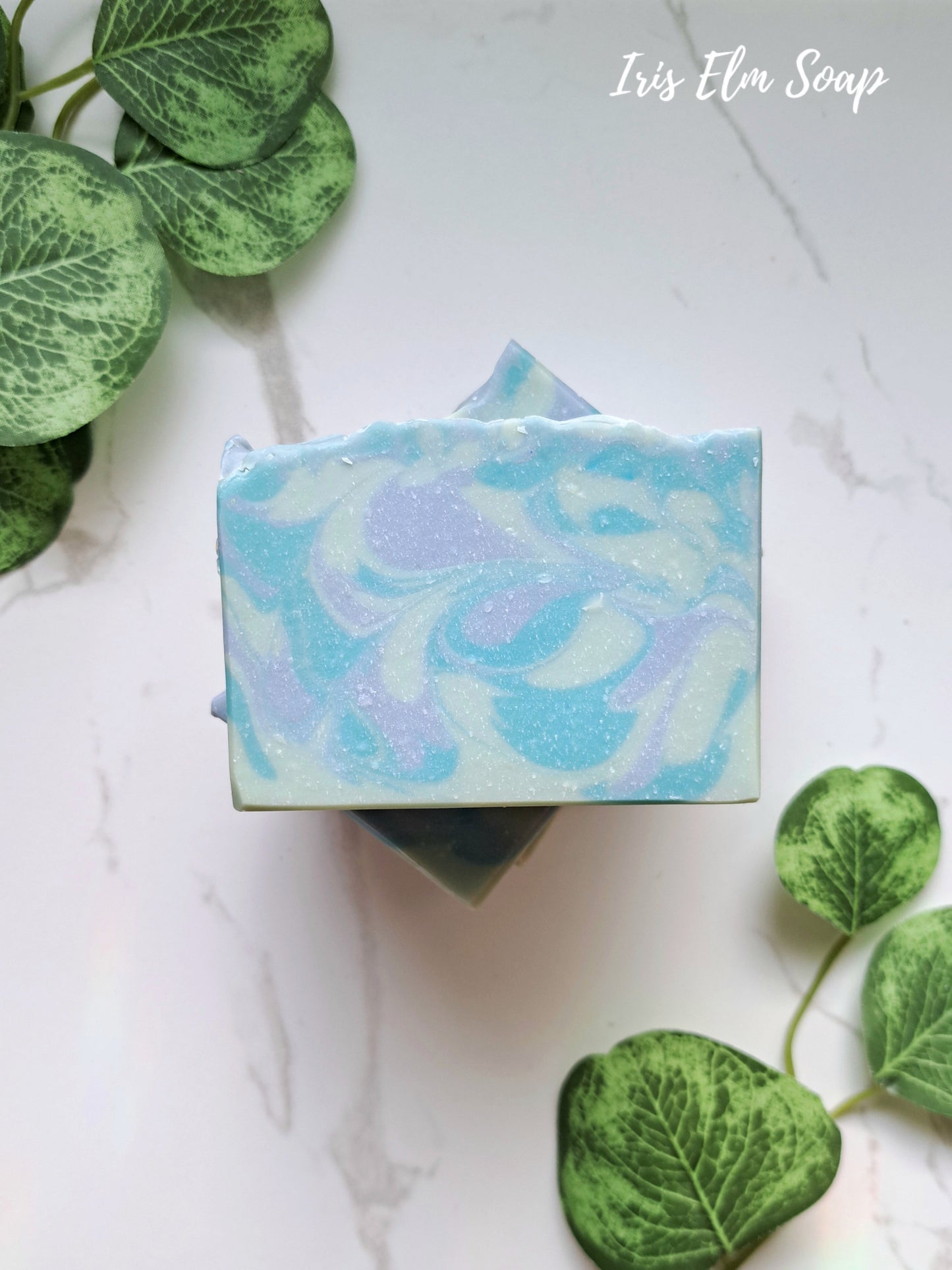 Marbled blue blueberry handmade soap bar with 'Iris Elm Soap' branding on a white background with green leaves.