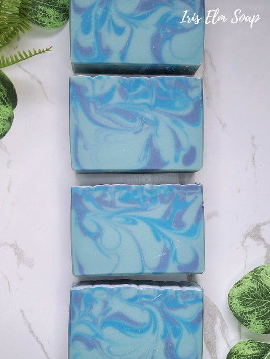Four rectangular bars of swirled blue blueberry handmade soap arranged vertically on a white marble surface, with green leaves placed around the edges. The text 'Iris Elm Soap' appears in the top right corner.