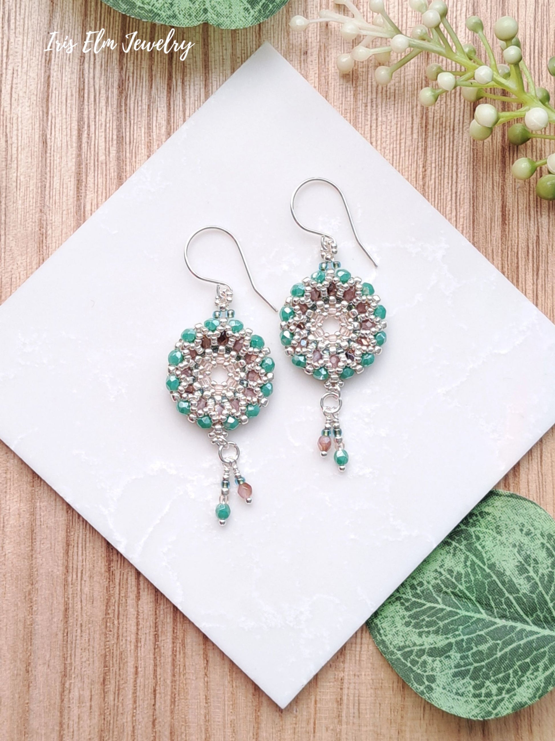 Pair of beaded turquoise blue and pink Czech glass woven wreath hoop earrings on a white card with green leaves and flowers in the background