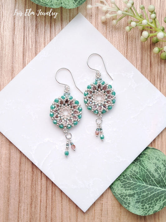 Pair of beaded turquoise blue and pink Czech glass woven wreath hoop earrings on a white card with green leaves and flowers in the background