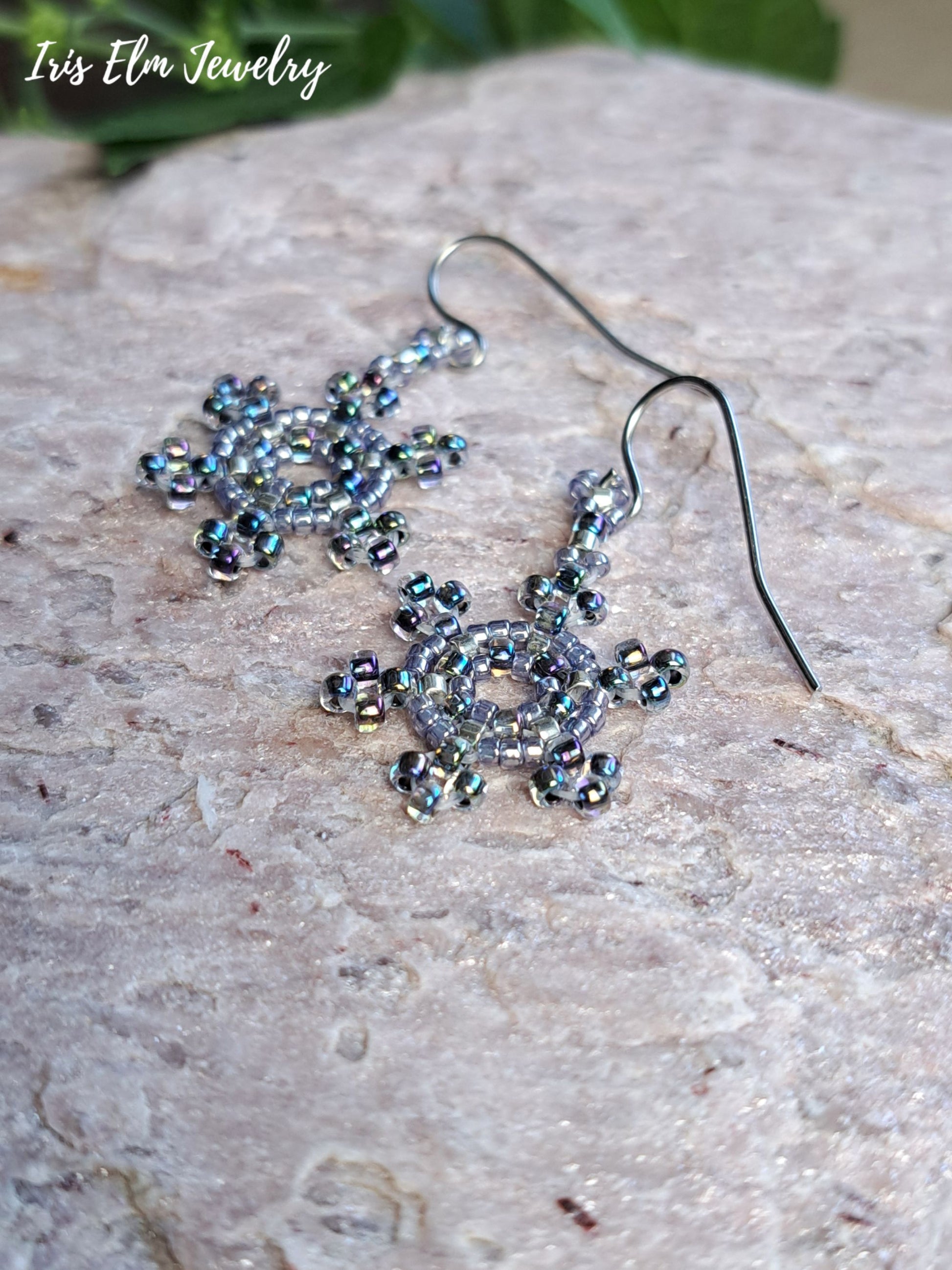 A pair of handmade snowflake-shaped dangle earrings made from iridescent silver beads on dark metal fishhook wires, displayed on a textured light beige stone surface with a green leaf in the background. The brand name 'Iris Elm Jewelry' is visible in the top left corner.