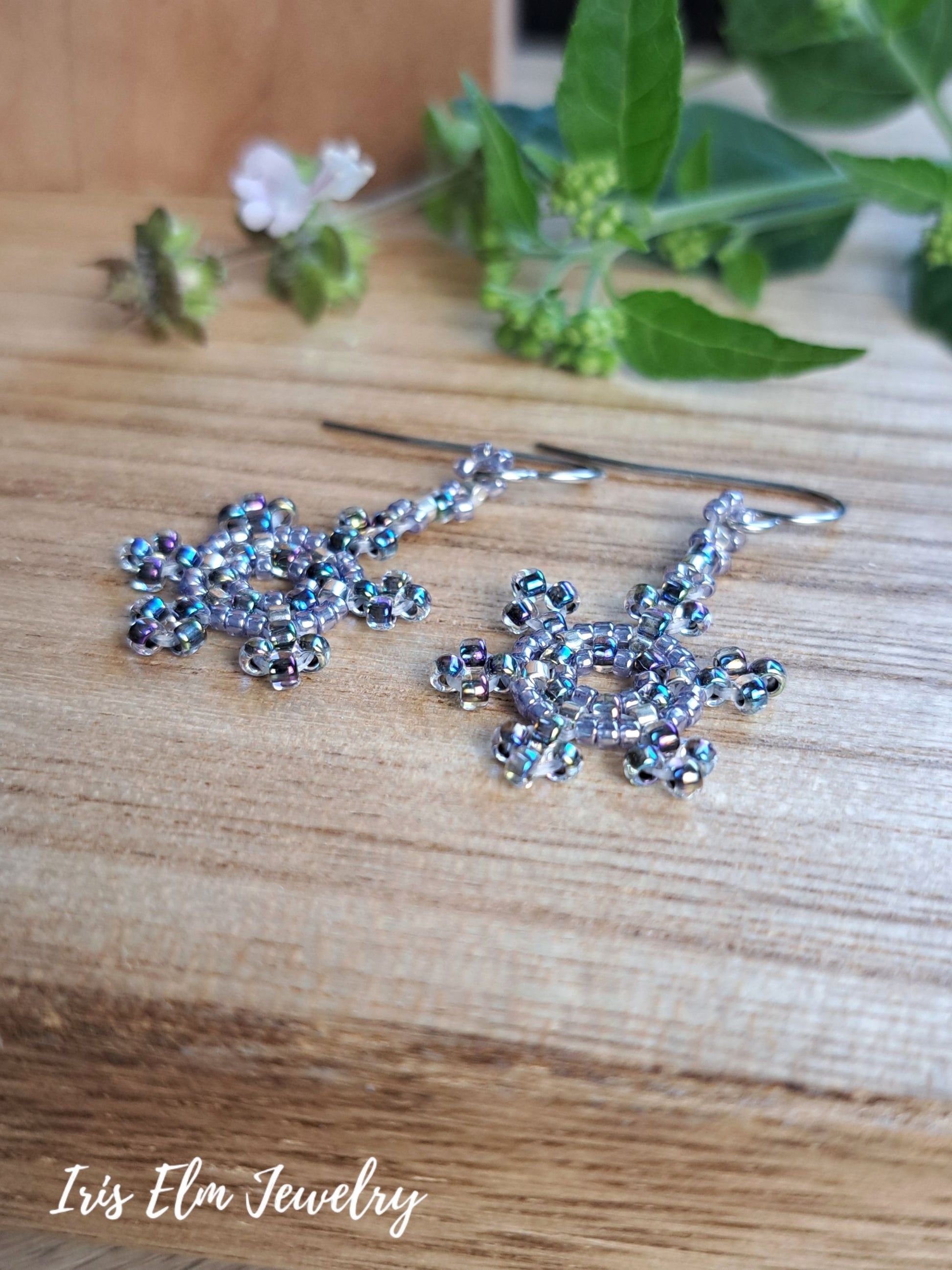 A pair of handmade beaded snowflake earrings with iridescent blue and silver beads on silver hooks, displayed on a wooden surface with green leaves in the background. The brand name 'Iris Elm Jewelry' is visible in the bottom-left corner.
