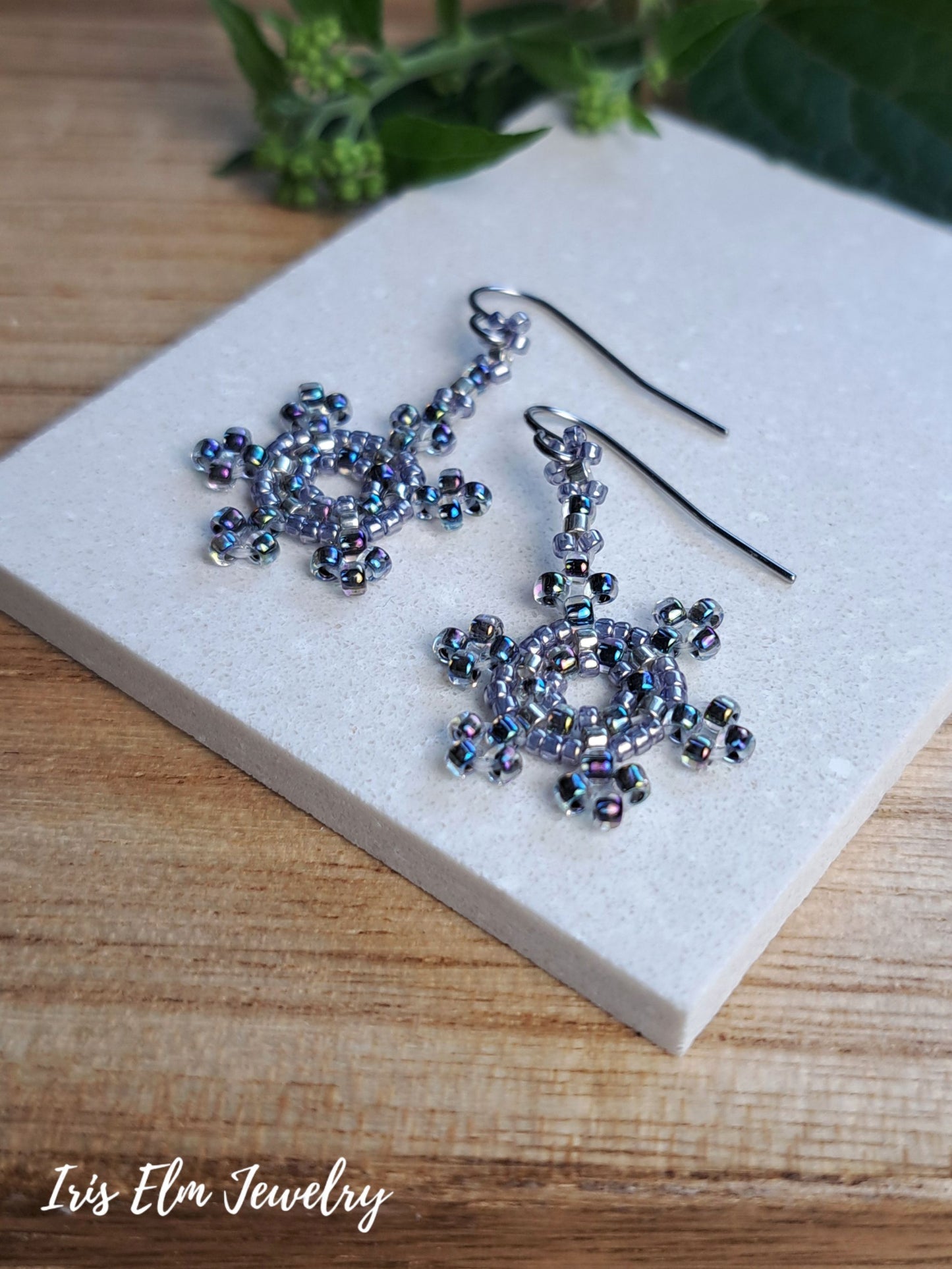 A pair of snowflake-shaped beaded dangle earrings with dark metal fishhooks, displayed on a light gray stone tile over a wooden surface. The beads are iridescent with a blue-purple shift.