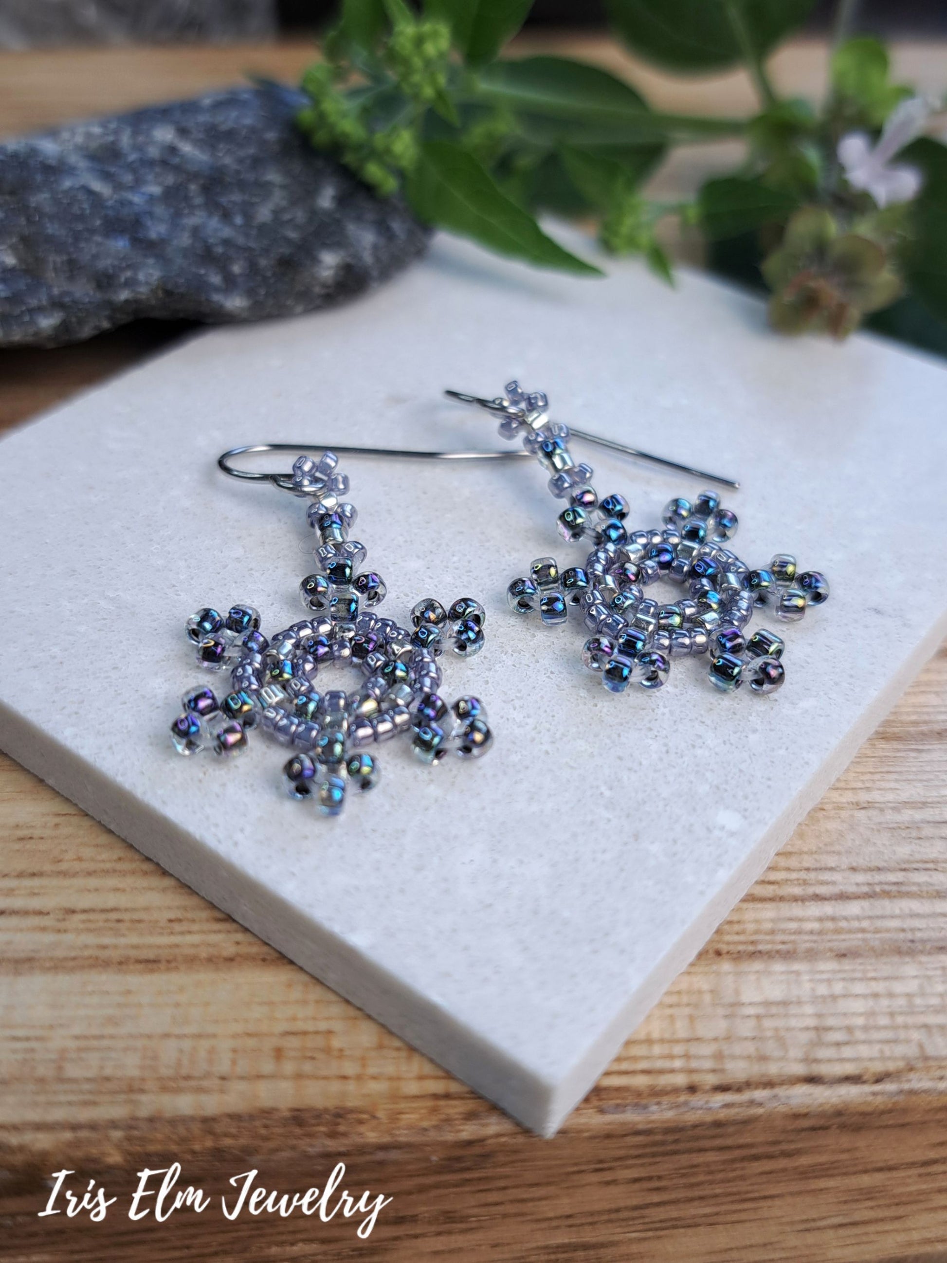 A pair of handmade beaded snowflake dangle earrings with iridescent blue and silver beads, displayed on a light gray stone slab over a wooden surface with green leaves in the background. The brand name 'Iris Elm Jewelry' is visible in the bottom-left corner.