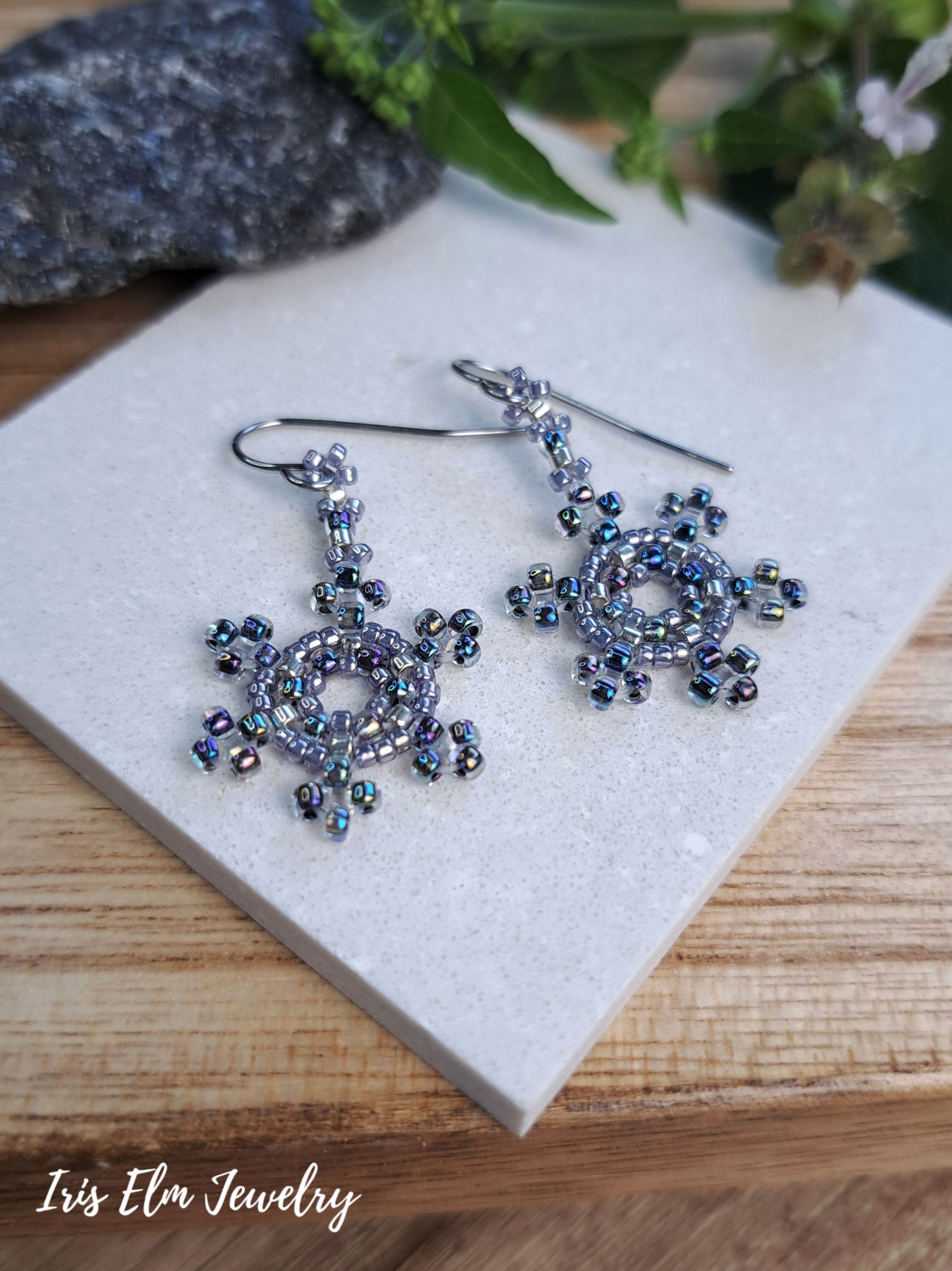 A pair of handmade beaded dangle earrings with silver-toned fishhook wires and intricate snowflake-like designs set with iridescent blue, purple, and green beads, displayed on a light gray stone slab over a wooden surface with the brand name 'Iris Elm Jewelry' in the bottom left corner.