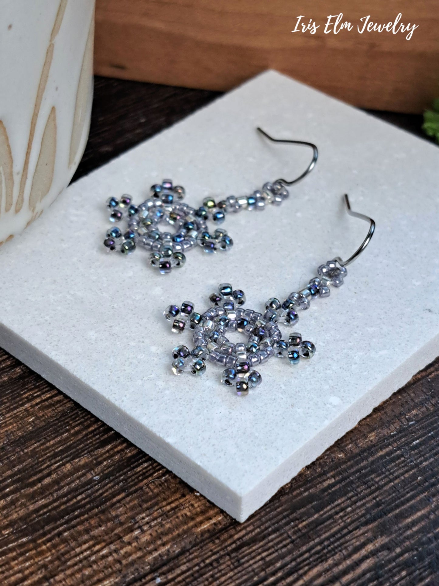 A pair of handmade beaded dangle earrings with silver-toned fishhook wires and iridescent blue-gray beads, displayed on a white textured tile over a dark wood surface. The brand name 'Iris Elm Jewelry' is visible in the top right corner.