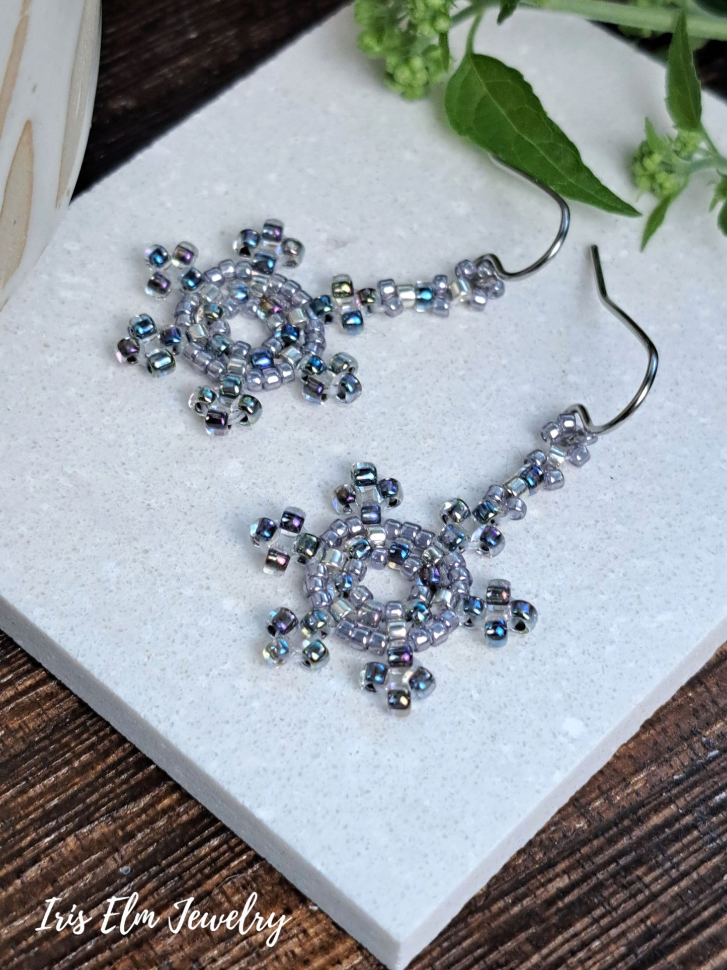 A pair of handmade beaded dangle earrings with silver-toned hooks and circular, snowflake-like beadwork in iridescent blue, purple, and silver, displayed on a white stone slab with green leaves in the background. The brand name 'Iris Elm Jewelry' is visible in the bottom-left corner.