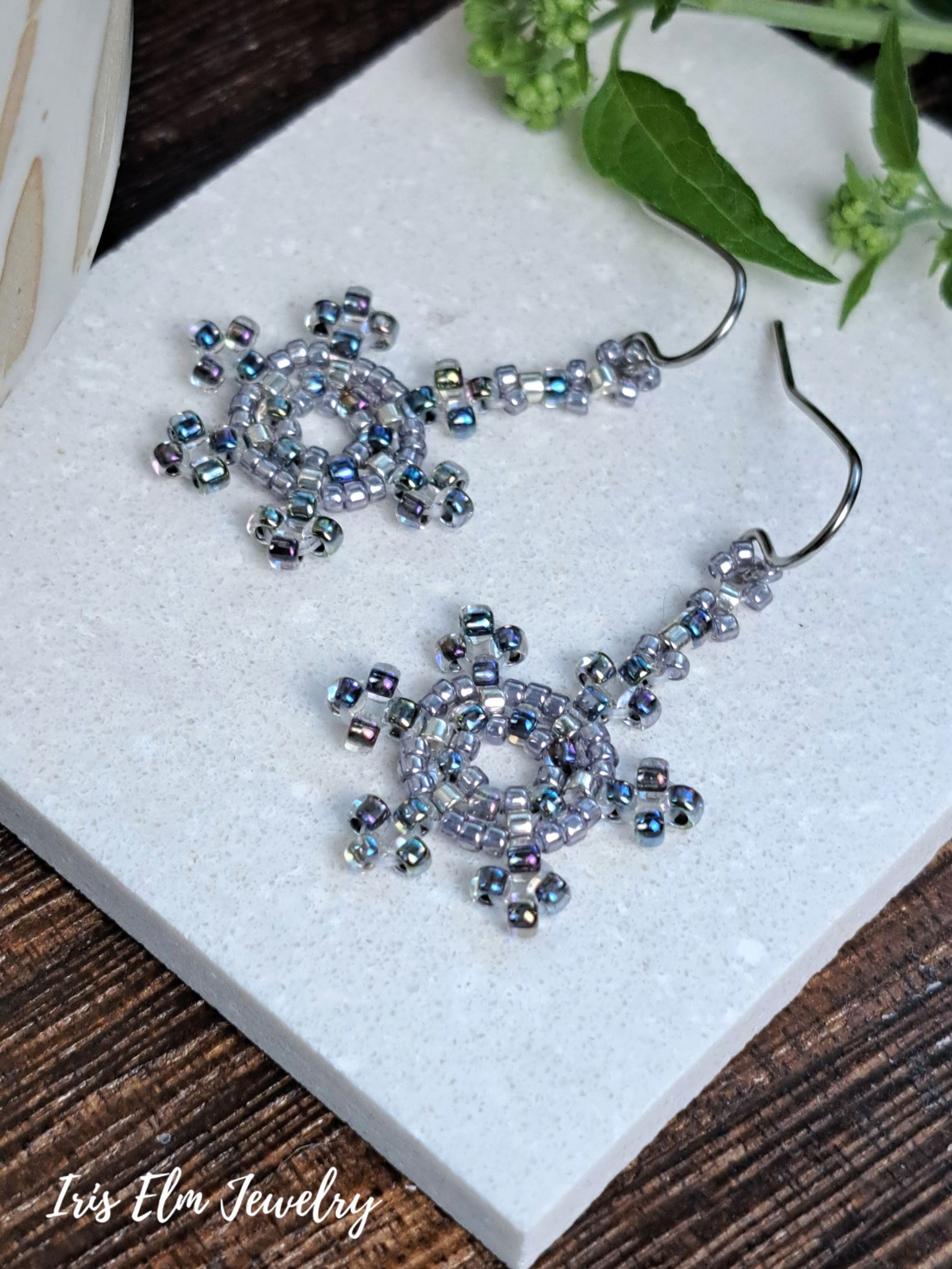 A pair of handmade beaded dangle earrings with silver-toned hooks and circular, snowflake-like beadwork in iridescent blue, purple, and silver, displayed on a white stone slab with green leaves in the background. The brand name 'Iris Elm Jewelry' is visible in the bottom-left corner.
