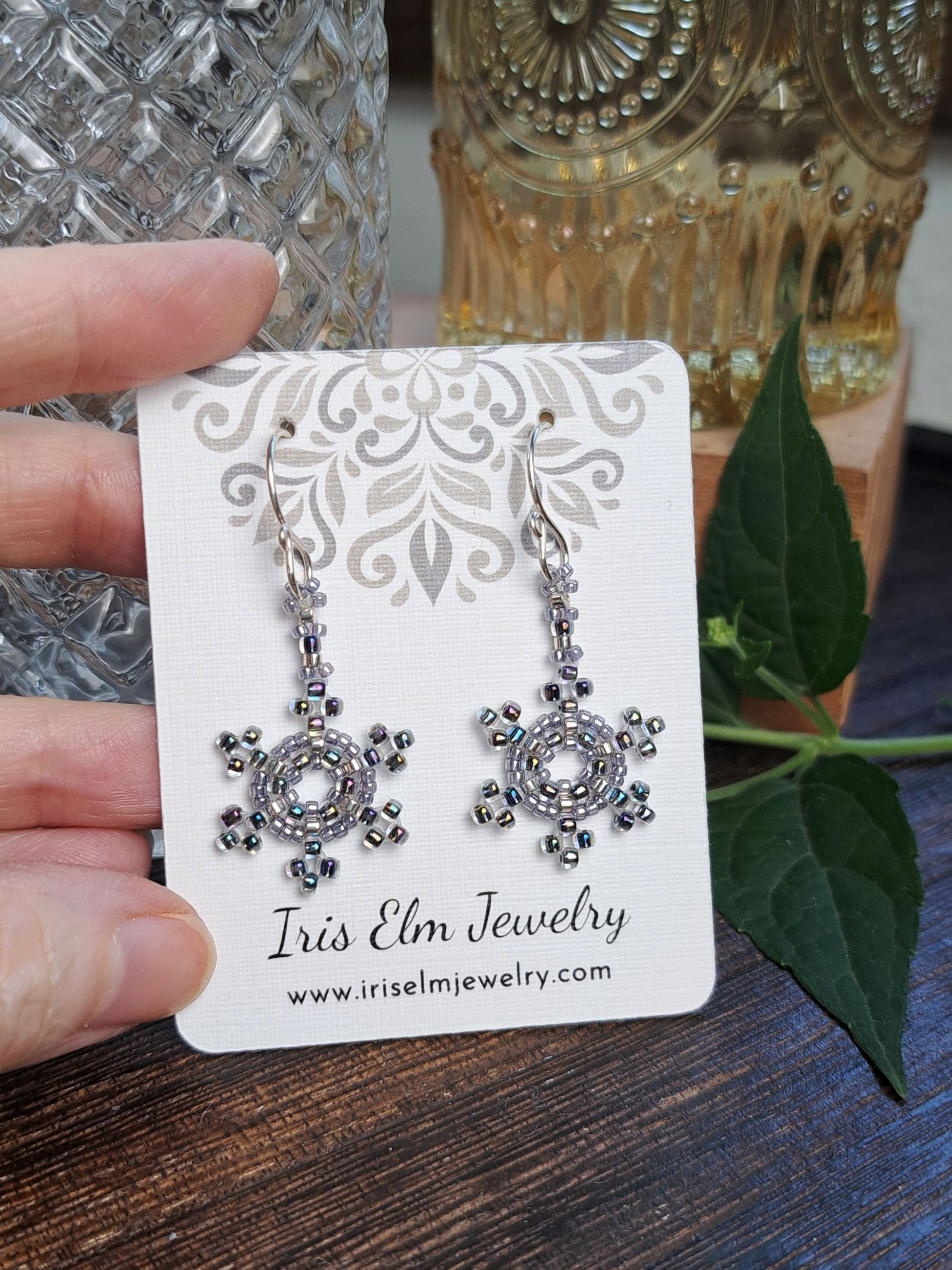 A hand holds a white jewelry card displaying a pair of dangling earrings with silver-toned metal and clear rhinestones in a circular sunburst design. The card reads 'Iris Elm Jewelry' with the website below.